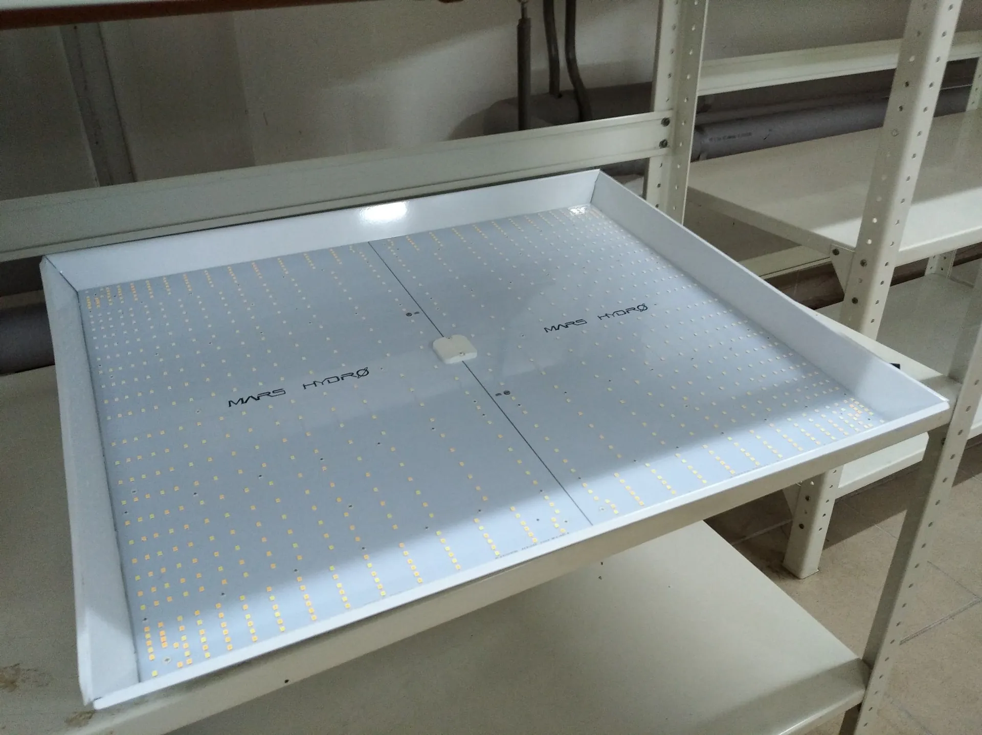 Biała, prostokątna lampa LED do uprawy roślin, leżąca na białym regale magazynowym. Powierzchnia lampy jest wypełniona gęstą siatką małych, żółtawych diod LED. Na obudowie widnieje napis 'MARS HYDRO' powtórzony w dwóch miejscach.