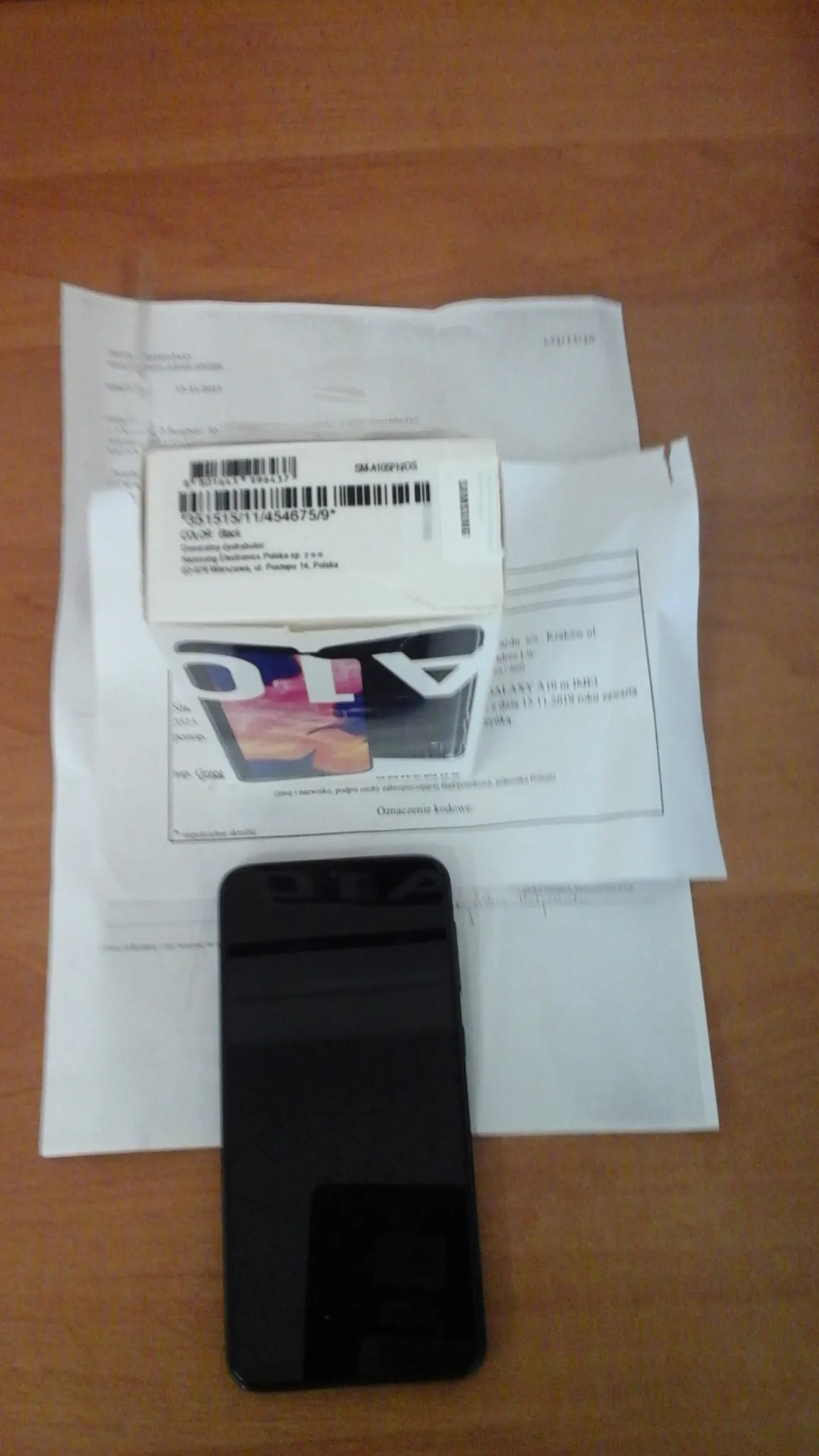 Na drewnianym stole leżą czarny smartfon, pudełko Samsung A10 z widocznym napisem 'A10' i kodami kreskowymi oraz kilka białych dokumentów z tekstem.