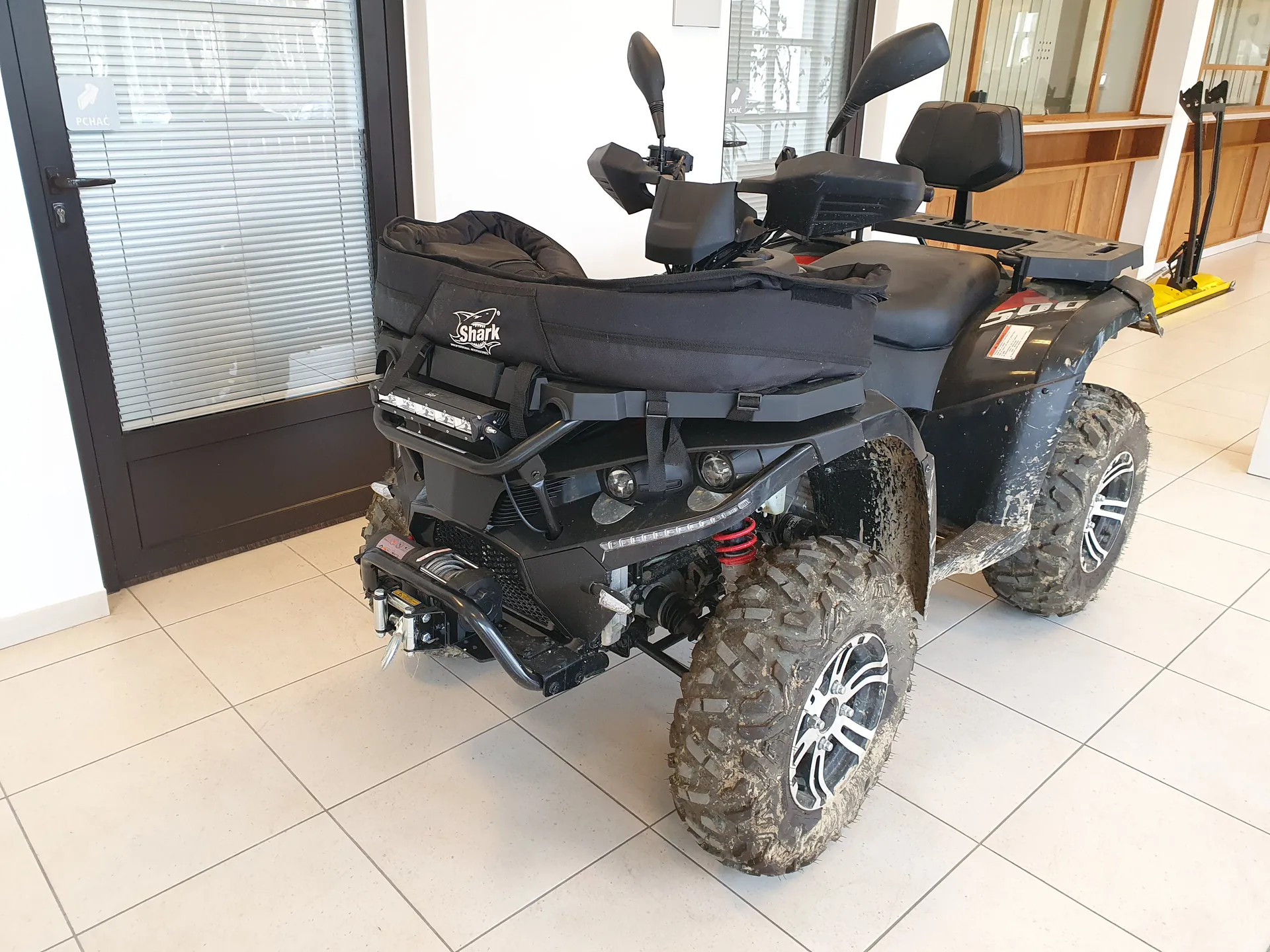 Czarny quad z oznaczeniem 500 i logo Shark na czarnej poduszce siedzeniowej, stojący w pomieszczeniu z białymi płytkami podłogowymi. Quad ma zabrudzone opony, czerwone elementy zawieszenia oraz czarne lusterka. W tle widoczne są drzwi z żaluzjami, drewniana szafka i żółty podnośnik. Na drzwiach znajduje się tabliczka z napisem PCHAĆ.