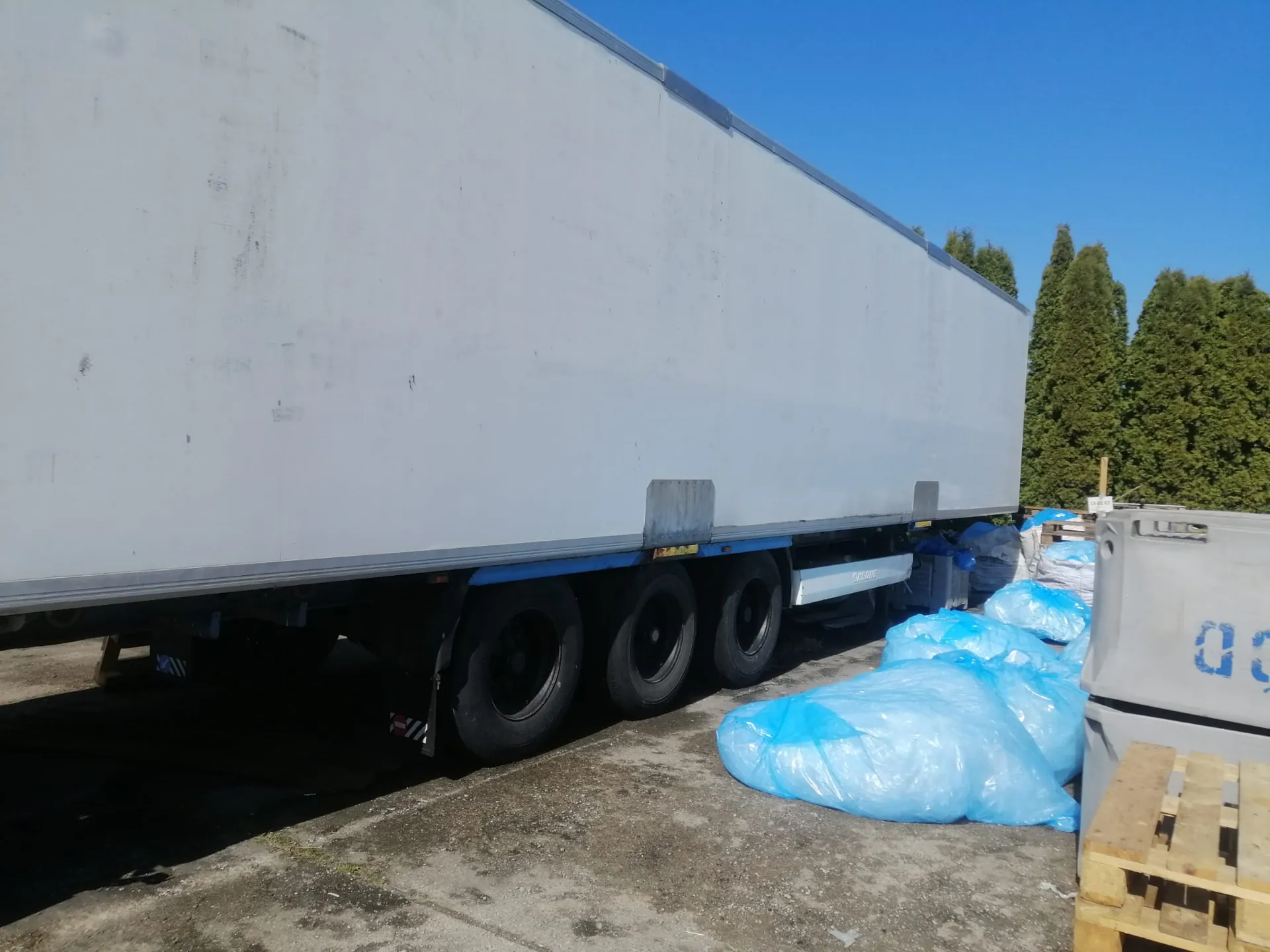 Boczny widok białej naczepy ciężarówki zaparkowanej na betonowym placu. Pod naczepą widoczne są trzy osie z kołami. Obok pojazdu leżą duże, niebieskie worki z tworzywa sztucznego oraz drewniana paleta i szary plastikowy pojemnik. W tle widać zielone drzewa i bezchmurne, niebieskie niebo.