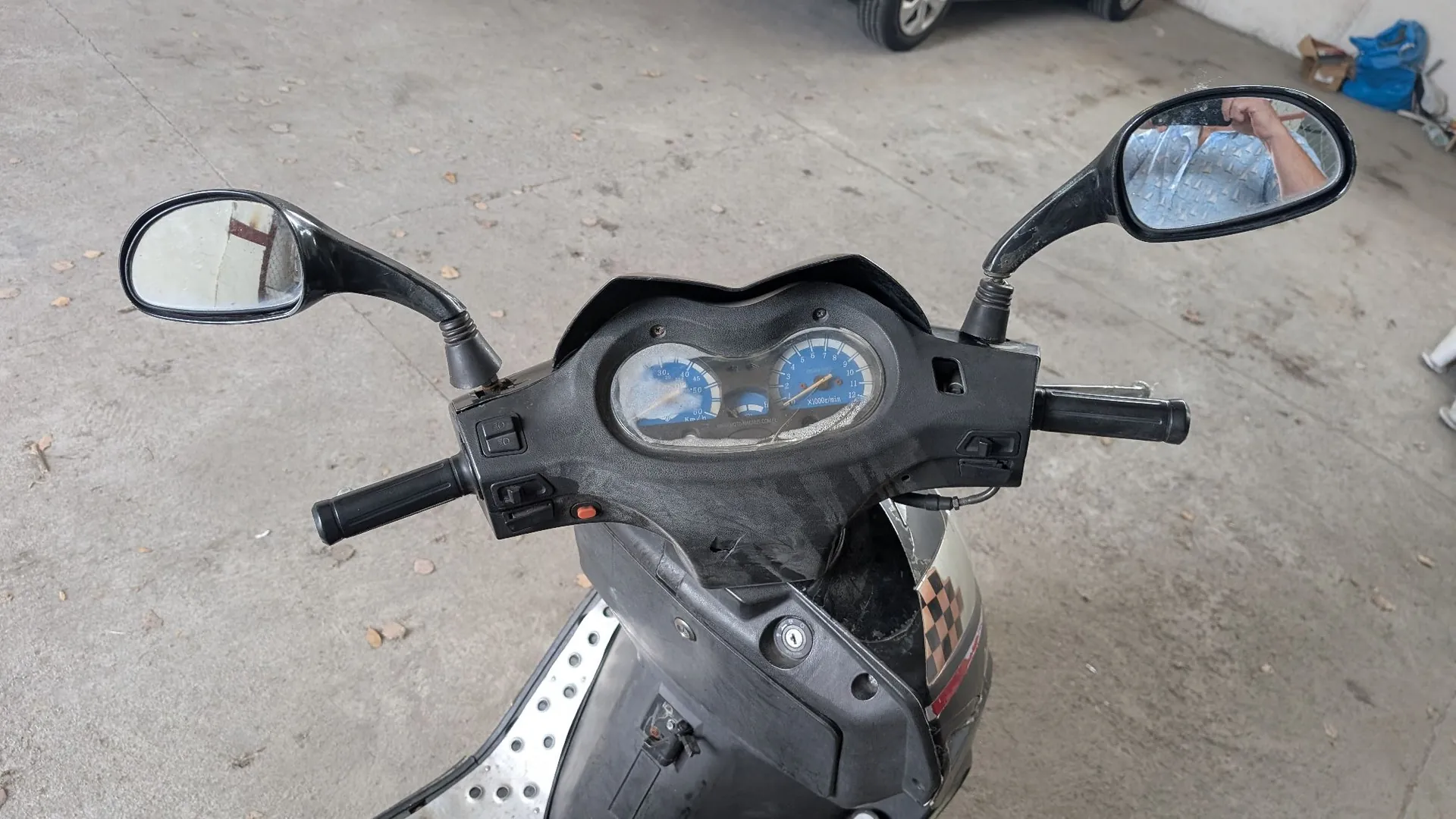 Widok z perspektywy kierowcy na kierownicę i deskę rozdzielczą czarnego skutera. Widoczne są dwa lusterka wsteczne, dwa wskaźniki analogowe (prędkościomierz i obrotomierz) oraz uchwyty kierownicy. Skuter posiada srebrne elementy z dziurkami na podnóżku i czarno-biały wzór szachownicy na przedniej obudowie. Pojazd stoi na betonowej posadzce.