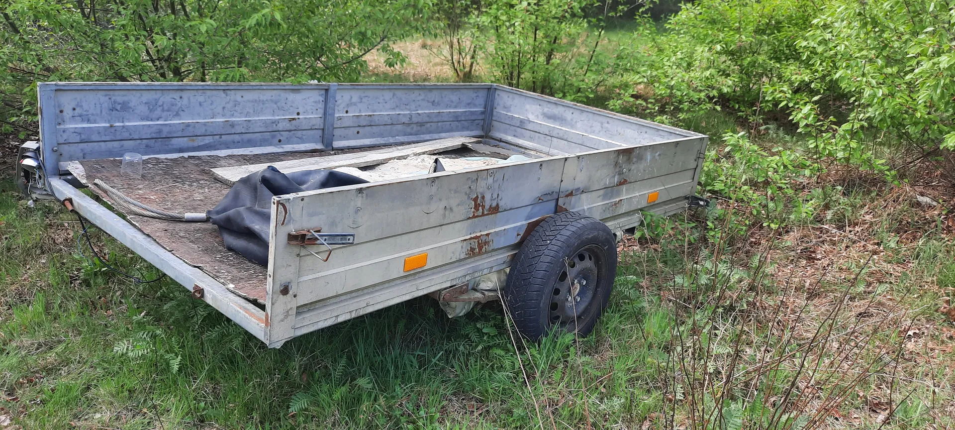 Stary metalowy przyczepa z widoczną rdzą, stojąca na trawie wśród zielonych krzewów i drzew. Wewnątrz przyczepy leży czarna folia, deski oraz szklany słoik. Przyczepa posiada jedno widoczne koło z oponą i pomarańczowe odblaski po bokach. Tło stanowi zarośla i trawa.
