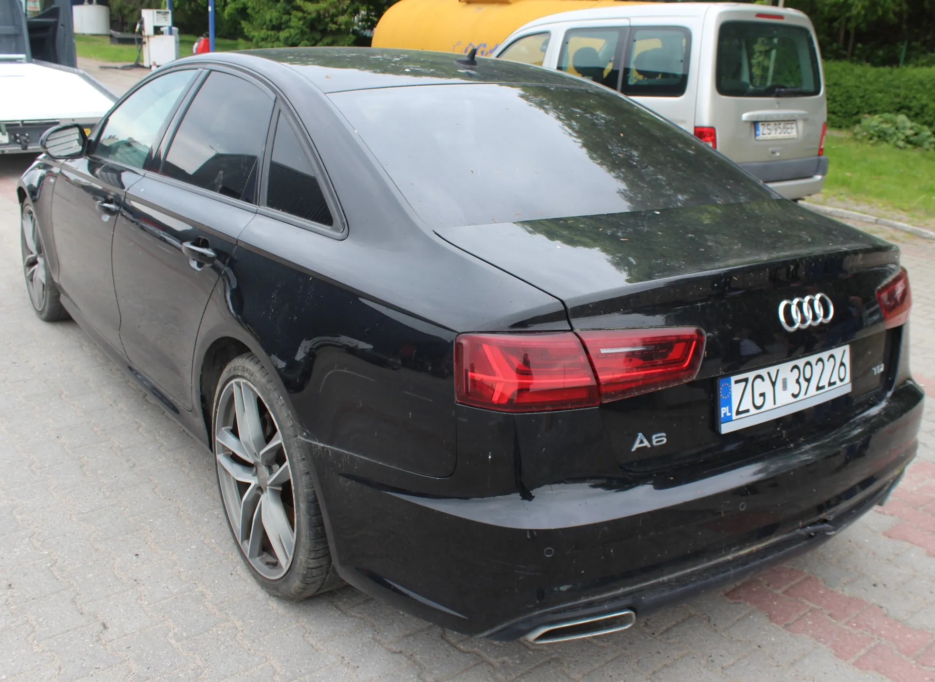 Czarny samochód marki Audi A6 zaparkowany na kostce brukowej, widok z tyłu z boku. Pojazd ma polską tablicę rejestracyjną o numerze ZGY 39226. W tle widoczny jest srebrny samochód dostawczy oraz żółty obiekt.