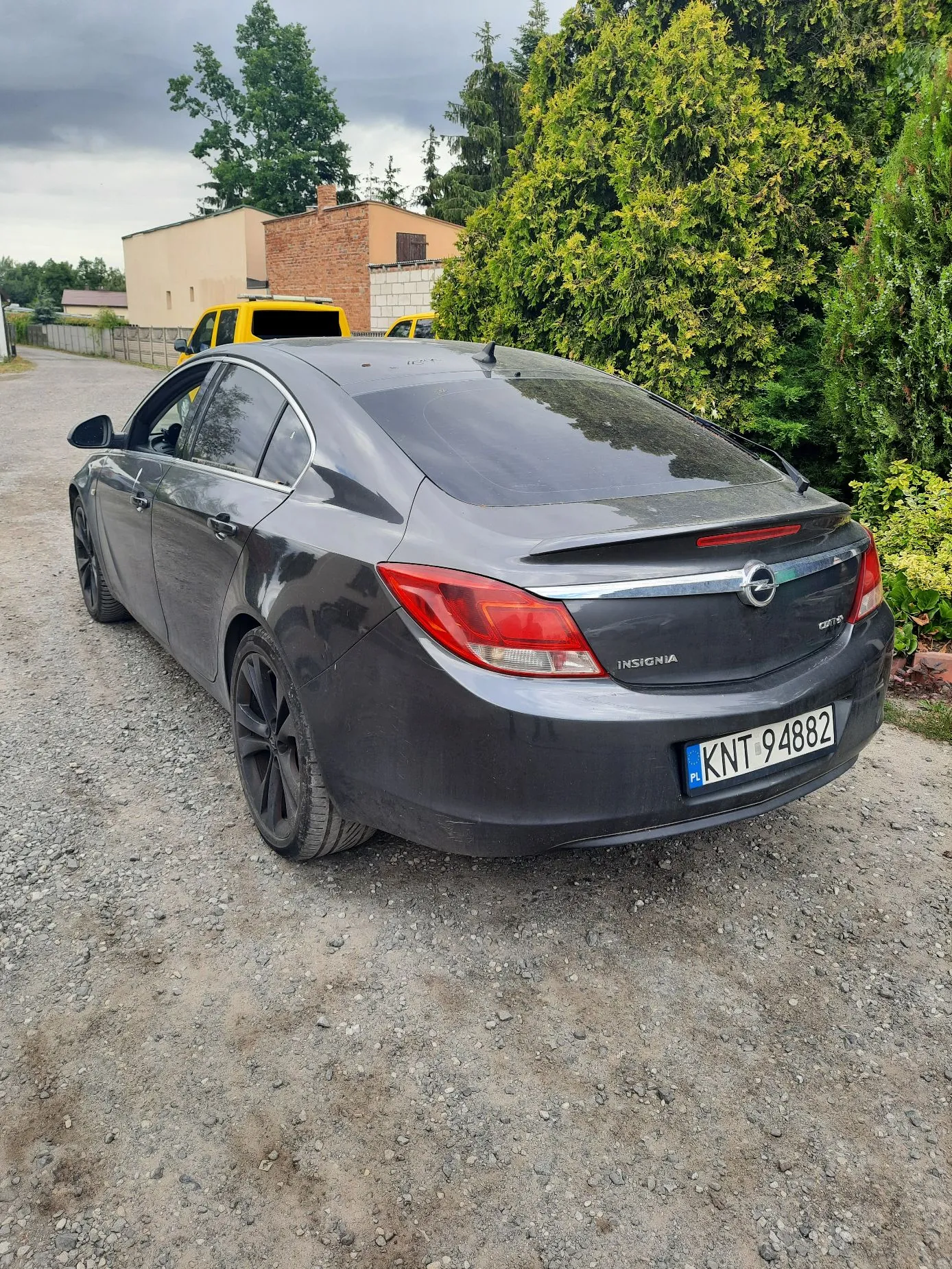 Szary samochód marki Opel Insignia zaparkowany tyłem na żwirowej drodze. Widoczna jest tylna część pojazdu z tablicą rejestracyjną o numerze KNT 94882. W tle znajduje się żółty samochód dostawczy, jasny budynek oraz gęste, zielone drzewa i krzewy pod zachmurzonym niebem.