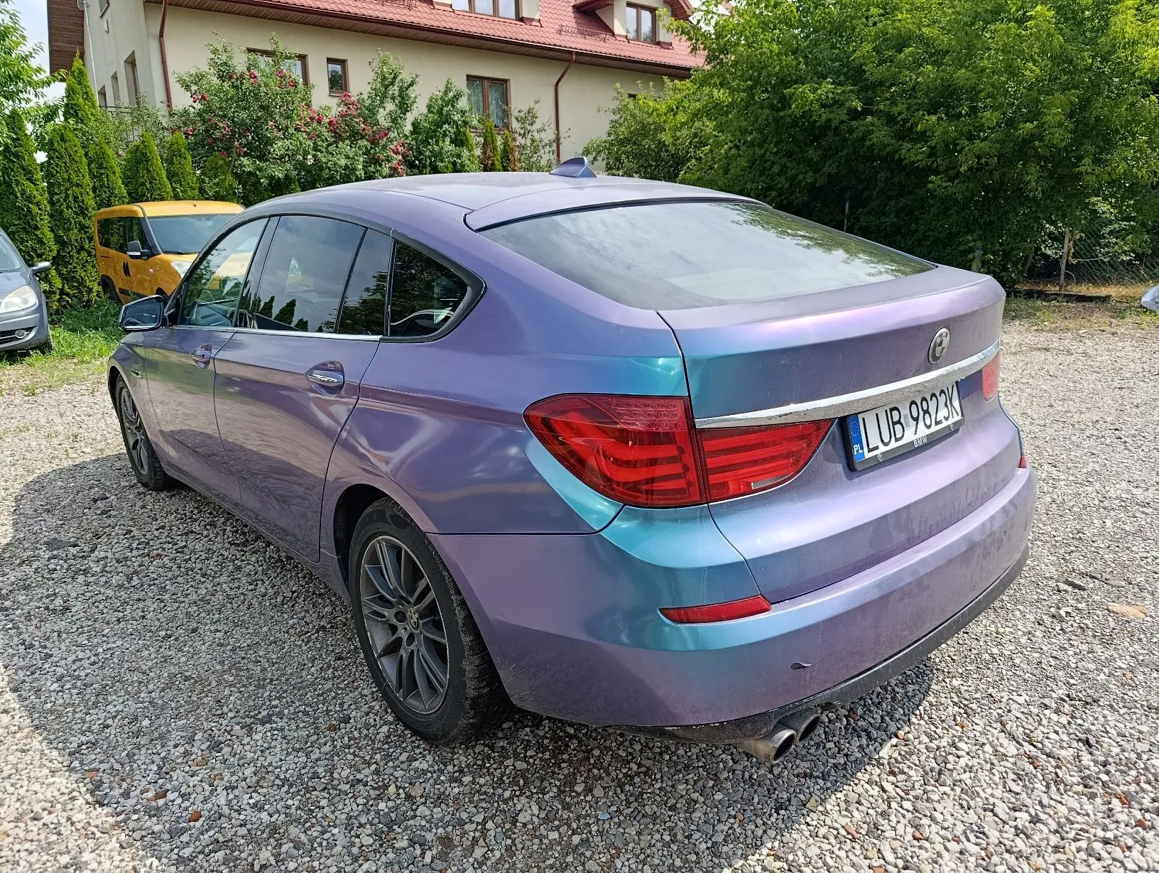 Tylny lewy profil samochodu marki BMW w kolorze opalizującym z fioletem i błękitem, zaparkowanego na żwirowym podjeździe. Pojazd posiada polską tablicę rejestracyjną o numerze LUB 9823K. W tle widoczny jest żółty samochód, zielone krzewy oraz fragment jasnego budynku z czerwonym dachem.