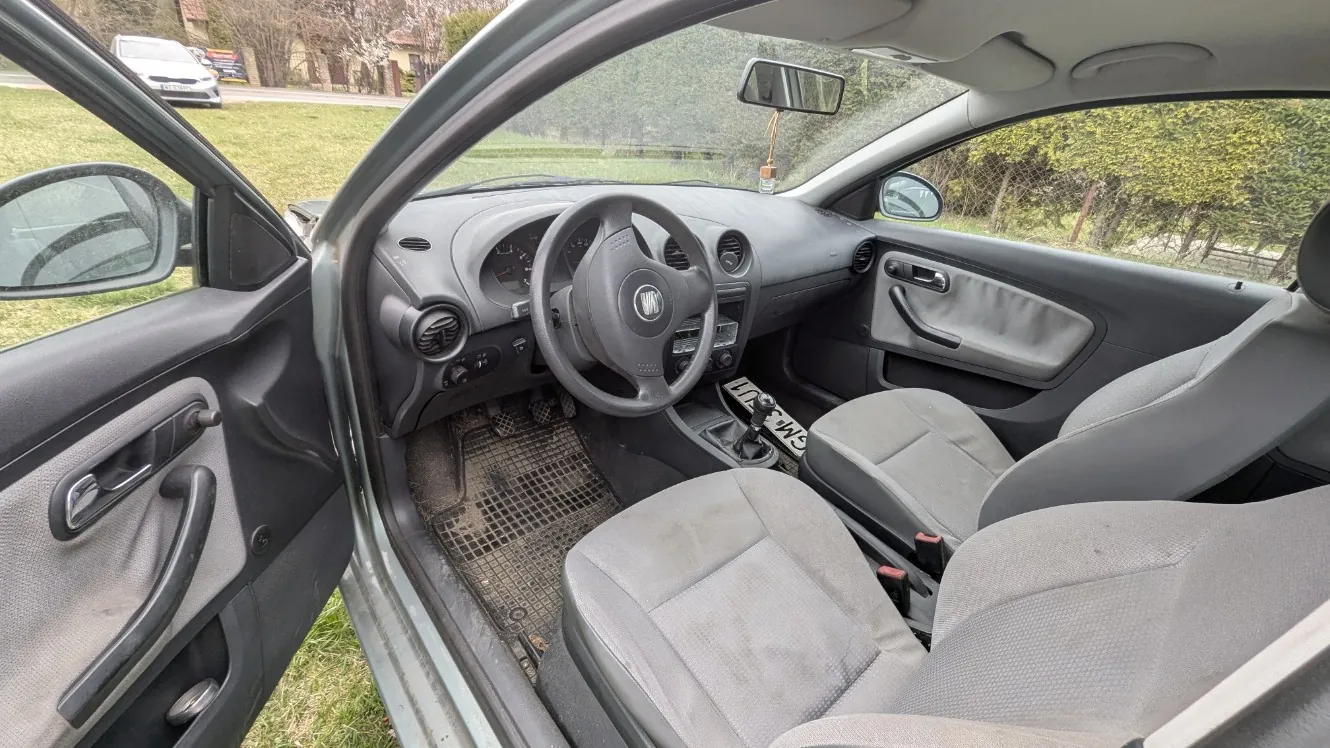 Wnętrze samochodu z otwartą lewą przodem drzwiami. Widoczne są szare siedzenia, kierownica z logo, deska rozdzielcza z licznikami, dźwignia zmiany biegów oraz dywaniki podłogowe. Na dywaniku w okolicy pedałów widoczny fragment napisu 'LIFES'. Poza samochodem trawa, droga oraz inne pojazdy na tle. W lusterku wstecznym i bocznym odbicia otoczenia.