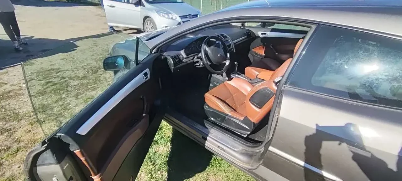 Otwarty bagażnik samochodu z widocznymi wewnętrznymi elementami, takimi jak przewody elektryczne, mechaniczna część oraz czarna podkładka z białą etykietą. Po lewej stronie widoczna część zewnętrzna samochodu z tylnym światłem. W tle trawa i światło słoneczne.