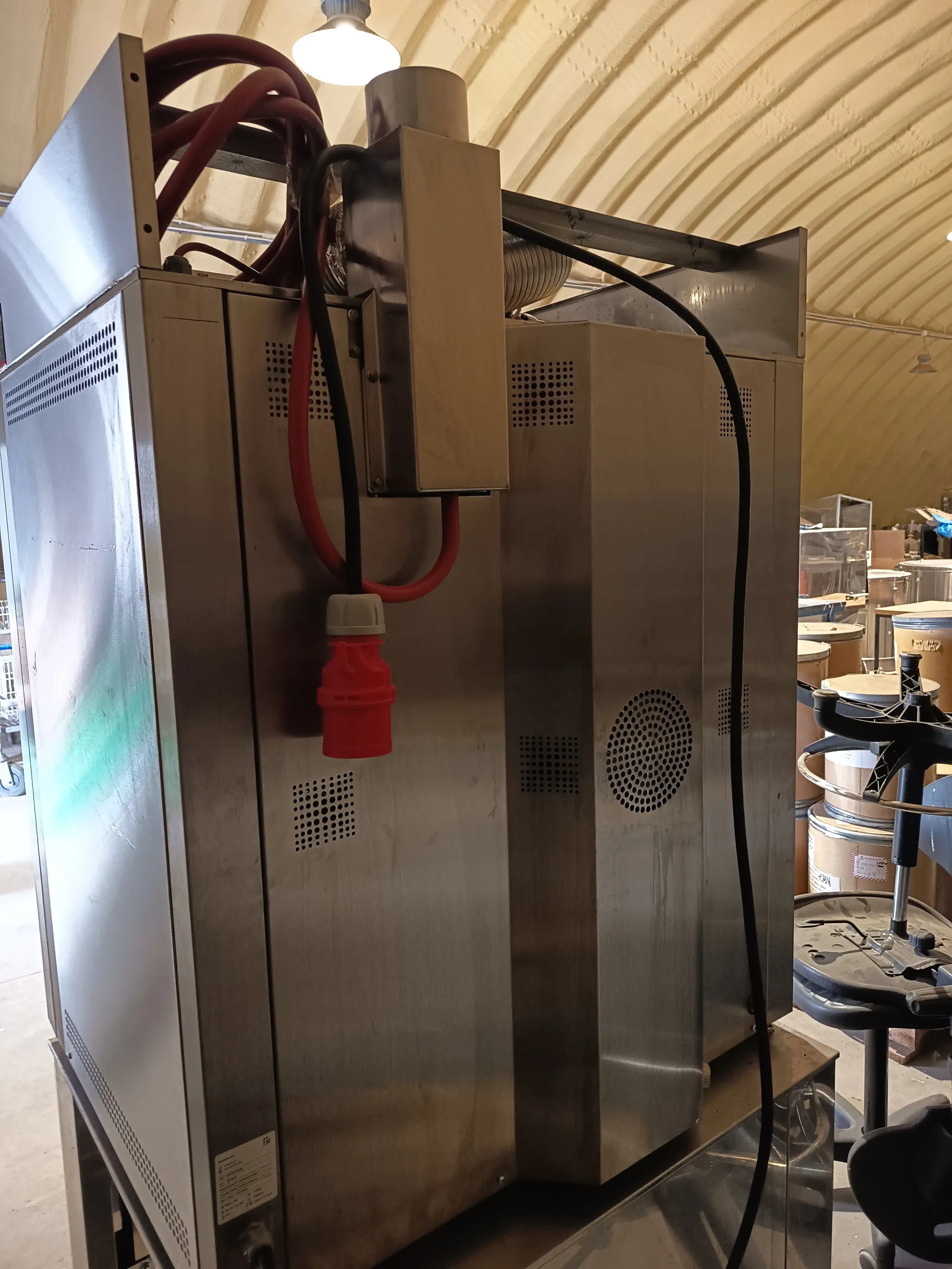 Wielki przemysłowy aparat ze stali nierdzewnej z czerwonymi i czarnymi przewodami oraz czerwonym wtyczkiem. Na maszynie widoczne są otwory wentylacyjne. Tło stanowi pomieszczenie z łukowym sufitem, oświetleniem oraz beczkami i innym sprzętem w oddali.