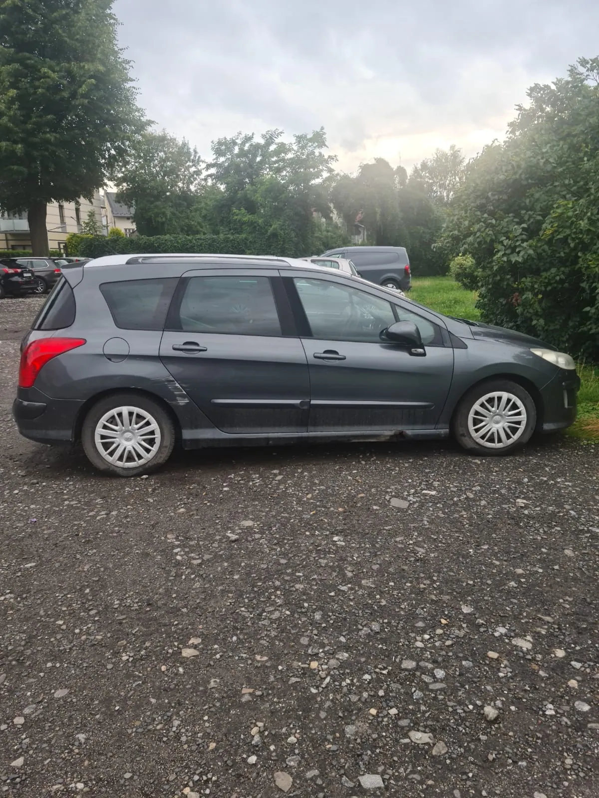 Ciemnoszary samochód typu kombi zaparkowany na żwirowym placu. Pojazd ma srebrne felgi i ciemne szyby. W tle widoczne są inne samochody, zielone krzewy, drzewa oraz budynki. Niebo jest zachmurzone. Na pierwszym planie znajduje się żwirowa powierzchnia z rozproszonymi kamieniami.