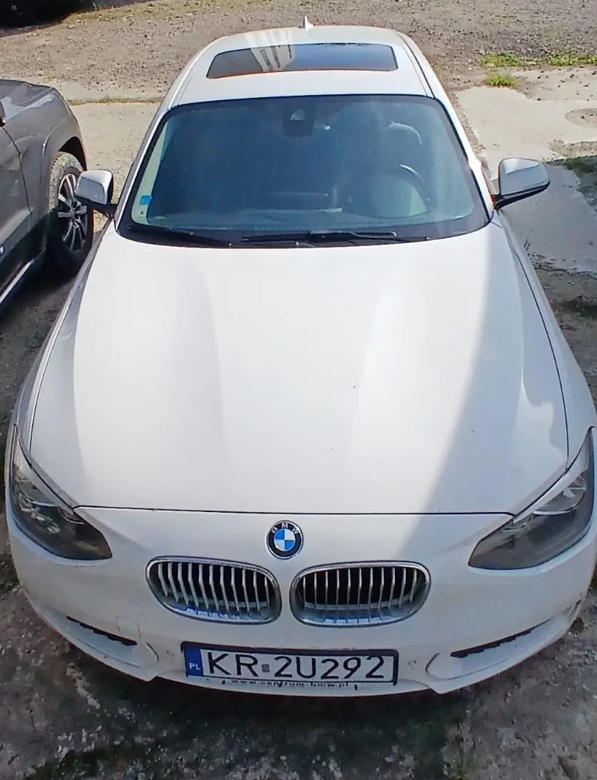 Widok z przodu na biały samochód marki BMW z panoramicznym dachem. Pojazd posiada polską tablicę rejestracyjną o numerze KR 2U292. Samochód zaparkowany jest na betonowym podłożu.