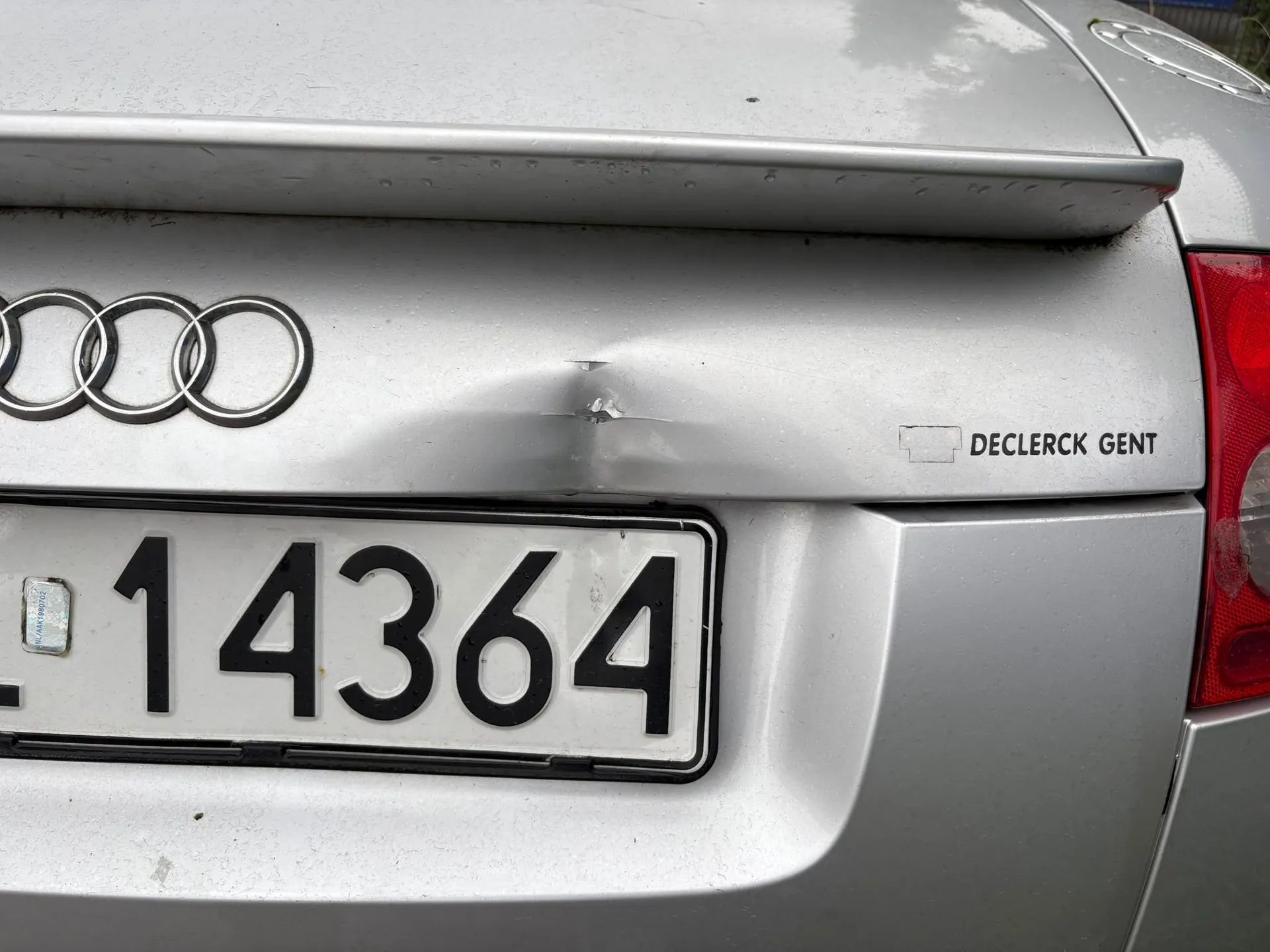 Zbliżenie na tylną część srebrnego samochodu marki Audi. Na klapie bagażnika widoczne jest logo z czterema splecionymi pierścieniami oraz wgniecenie z uszkodzeniem lakieru. Po prawej stronie znajduje się napis 'DECLERCK GENT'. Poniżej zamontowana jest tablica rejestracyjna z numerem '14364'. Po prawej stronie widoczny jest fragment czerwonego tylnego światła.