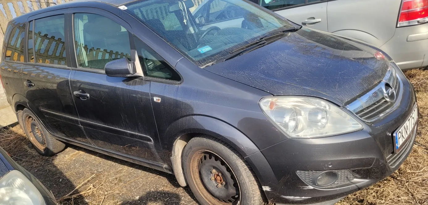 Ciemnoszary samochód osobowy marki Opel (model Zafira) zaparkowany na podwórku z suchą trawą i żwirem. Samochód ma zabrudzoną karoserię z widocznymi zadrapaniami, brakującą nakładki na felgach przedniego koła prawego oraz tablicę rejestracyjną z widocznym numerem PKN 777. W tle częściowo widoczny srebrny samochód i drewniany płot. Słońce oświetla scenę, rzucając cienie na ziemię.