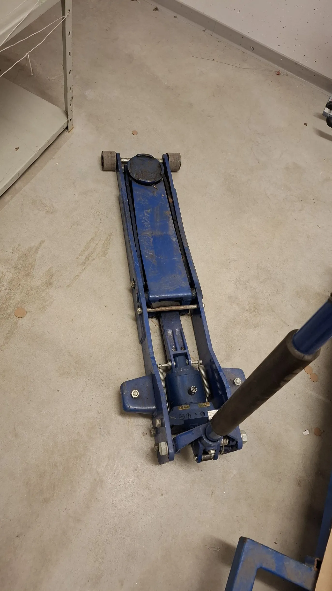 Niebieski podnośnik hydrauliczny typu żaba leżący na jasnoszarej, betonowej podłodze. Urządzenie jest widoczne z góry, z rączką do pompowania skierowaną w prawą stronę obrazu.