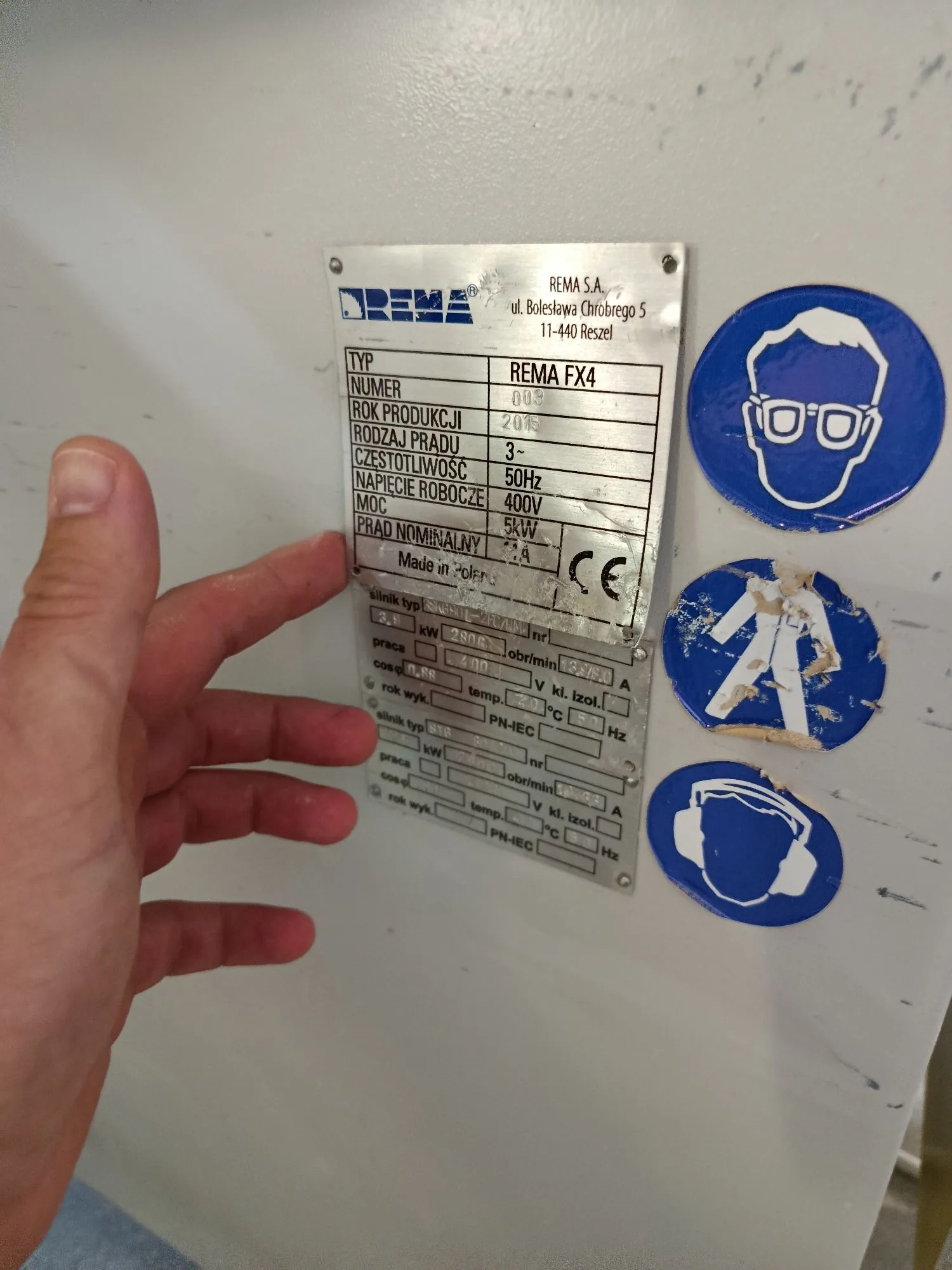 Zdjęcie tabliczki znamionowej urządzenia marki REMA S.A. z siedzibą w Reszlu. Na górnej tabliczce widnieją dane: TYP: REMA FX4, NUMER: 00, ROK PRODUKCJI: 2015, RODZAJ PRĄDU: 3~, CZĘSTOTLIWOŚĆ: 50Hz, NAPIĘCIE ROBOCZE: 400V, MOC: 5kW, PRĄD NOMINALNY: 11.4 A. Widoczne jest oznaczenie CE. Poniżej znajduje się druga, bardziej zużyta tabliczka z danymi technicznymi silnika. Po prawej stronie tabliczek znajdują się trzy niebieskie piktogramy BHP: obowiązek stosowania okularów ochronnych, znak ostrzegawczy z sylwetką człowieka oraz obowiązek stosowania ochrony słuchu. Po lewej stronie zdjęcia widoczna jest dłoń osoby wskazująca na tabliczkę.