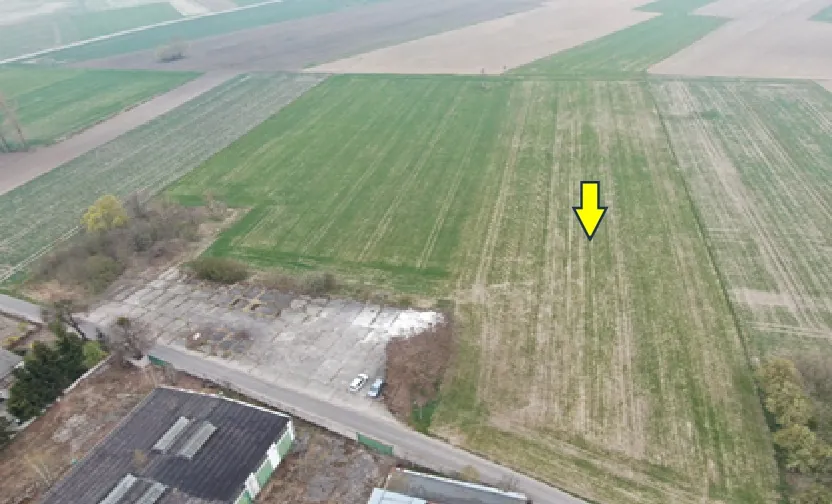 Zdjęcie lotnicze przedstawiające obszar rolniczy z polami w różnych odcieniach zieleni i brązu. W dolnej części obrazu widoczny jest utwardzony plac z zaparkowanymi samochodami oraz fragmenty budynków przemysłowych. Na jednym z zielonych pól znajduje się żółta strzałka skierowana w dół, wskazująca konkretny punkt na gruncie.