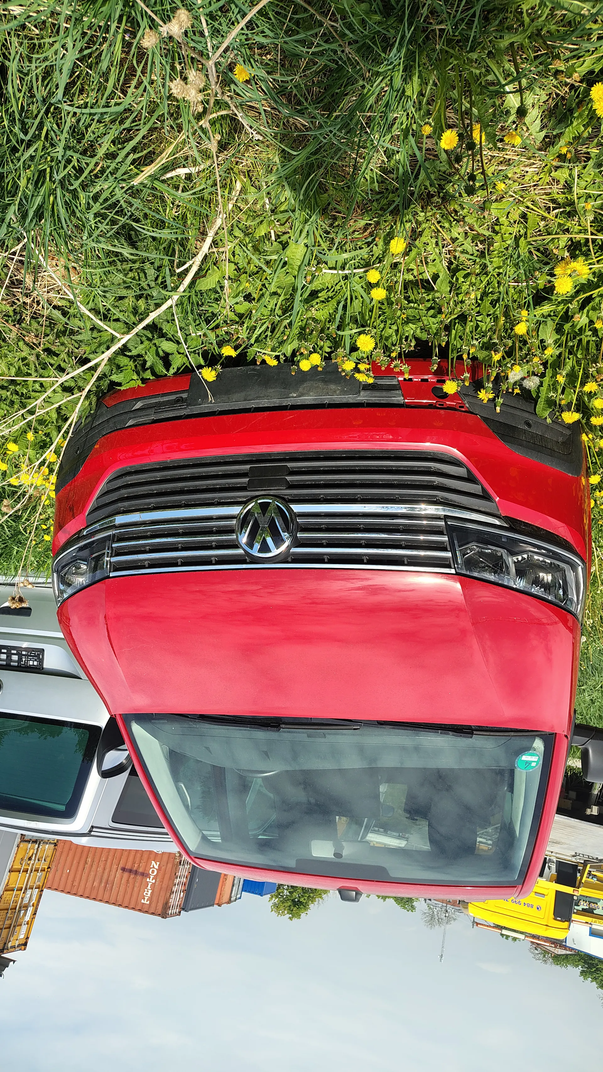 Czerwony samochód marki Volkswagen z przodem skierowanym w prawo, otoczony trawą z żółtymi kwiatami. W tle widoczny srebrny samochód, kontenery (jeden z napisem 'ZOP'), żółta konstrukcja oraz częściowo zachmurzone niebo.