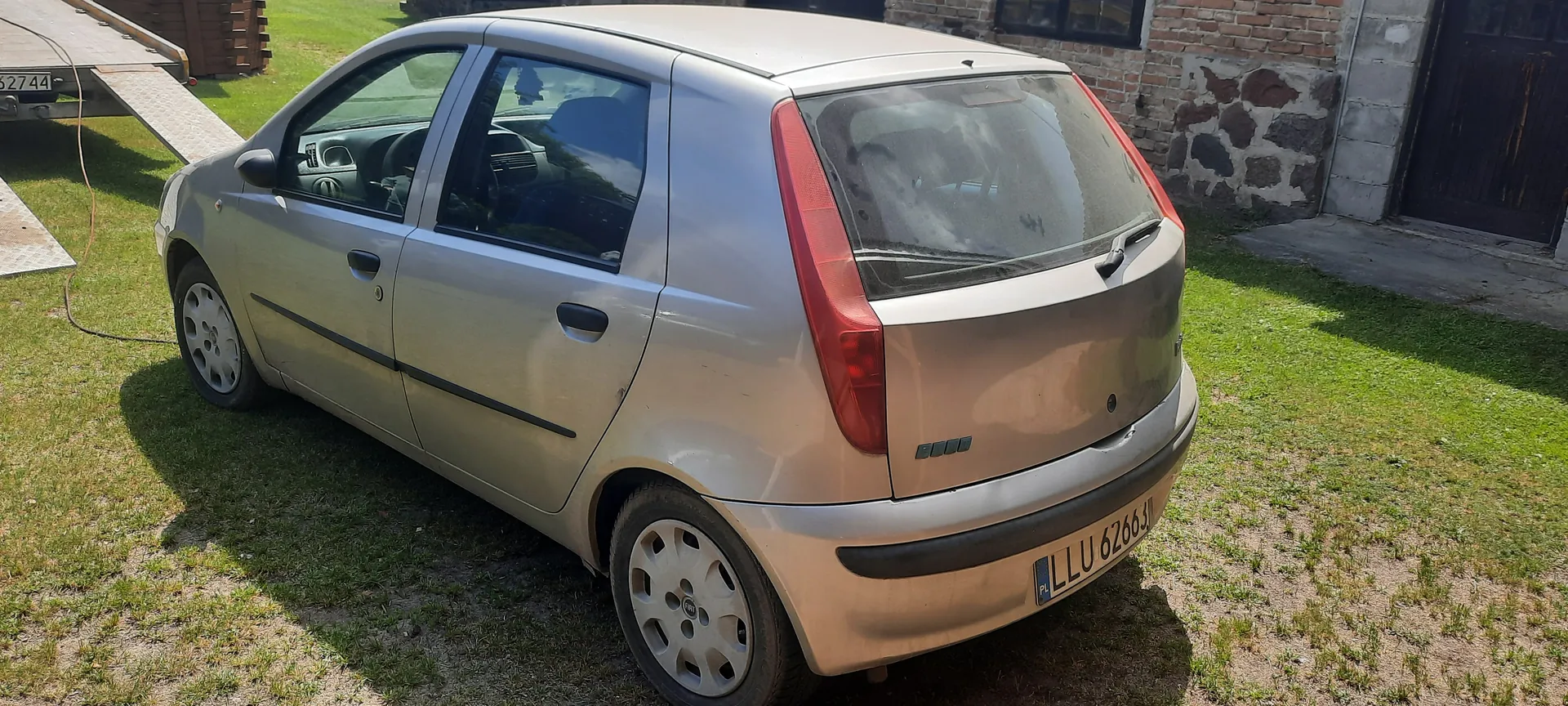 Srebrny samochód Fiat z tablicą rejestracyjną PL LLU 6266, zaparkowany na trawie. W tle ceglany budynek z kamieniem, ciemne drzwi i przyczepa z widocznym numerem 62744. Wnętrze samochodu jest częściowo widoczne przez szyby. Trawa na podwórku jest zielona z miejscami suchymi.