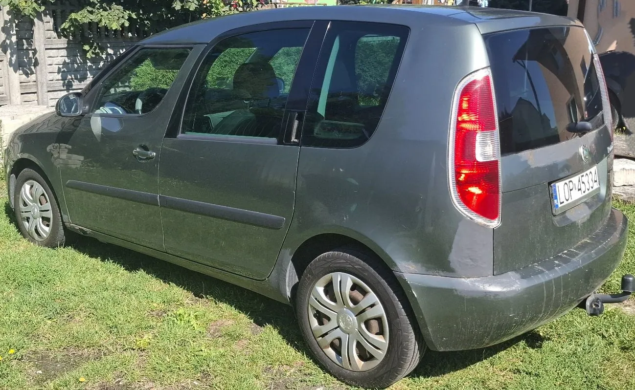 Szary samochód osobowy typu hatchback zaparkowany na trawie. Widok z boku i z tyłu. Pojazd posiada polską tablicę rejestracyjną o numerze LOP 45334 oraz hak holowniczy. W tle widoczny jest drewniany płot i zieleń.