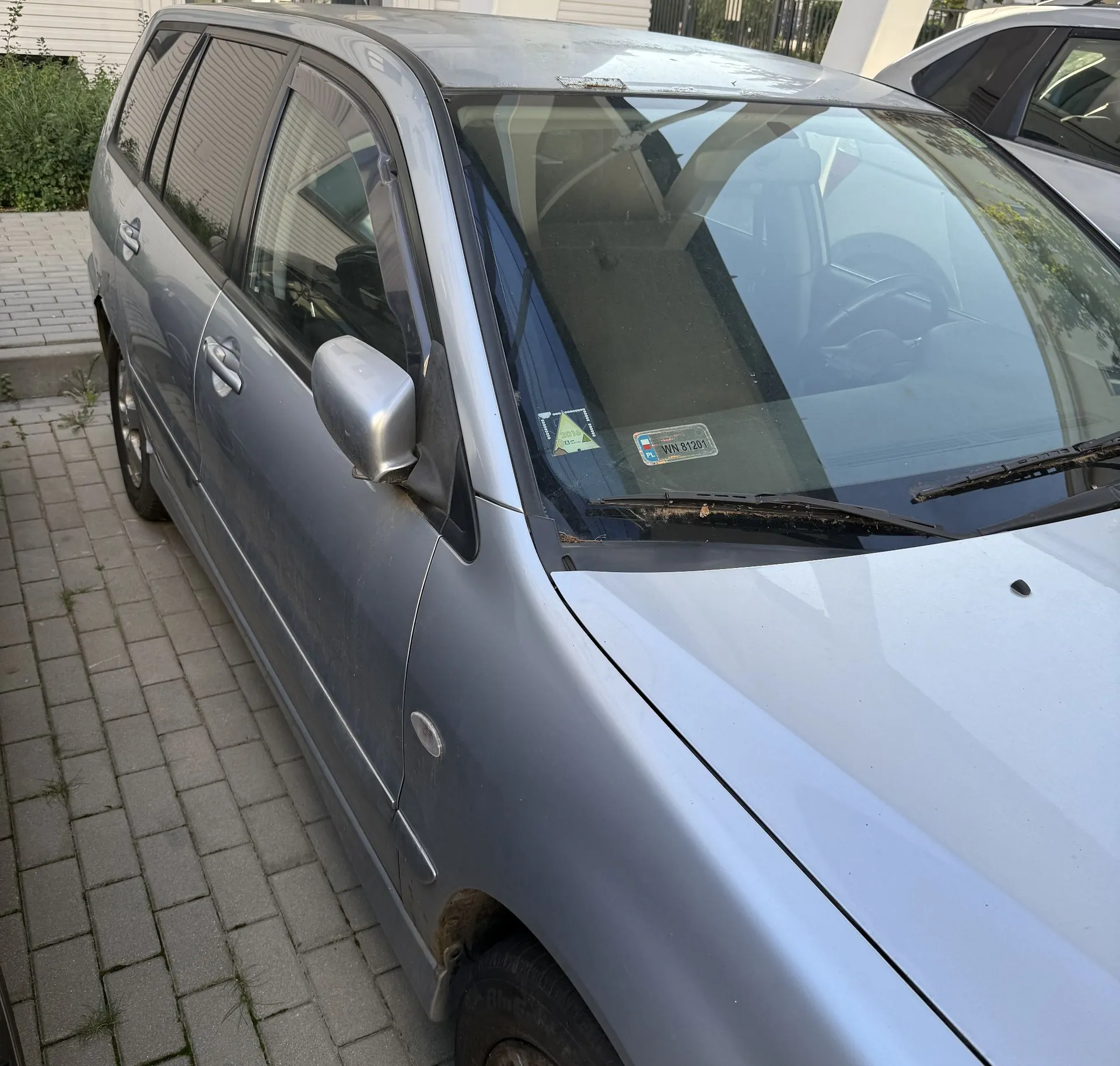 Srebrny samochód typu kombi zaparkowany na kostce brukowej. Widoczny jest przód i bok pojazdu, w tym przednia szyba z naklejką przeglądu technicznego o numerze WN 81201, lusterko boczne oraz przednie koło.