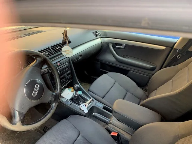 Wnętrze samochodu marki Audi widziane z perspektywy kierowcy. Widoczna jest czarna kierownica z logo czterech pierścieni, deska rozdzielcza, konsola środkowa z lewarkiem zmiany biegów oraz dwa przednie, ciemnoszare fotele materiałowe. Na lusterku wstecznym wisi okrągły zapach samochodowy. Na konsoli środkowej leżą kluczyki i drobne przedmioty.