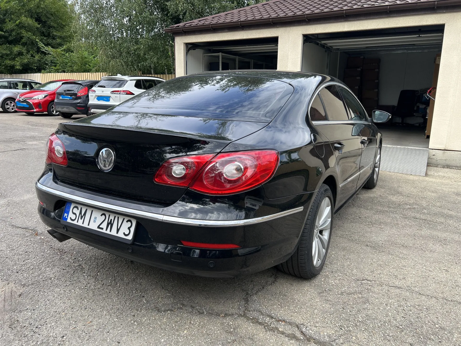 Czarny samochód marki Volkswagen widoczny od tyłu i z boku, zaparkowany na asfaltowym podjeździe przed garażem. Samochód ma polską tablicę rejestracyjną o numerze SMI 2WV3. W tle widoczne są inne zaparkowane samochody oraz budynek z otwartym garażem.