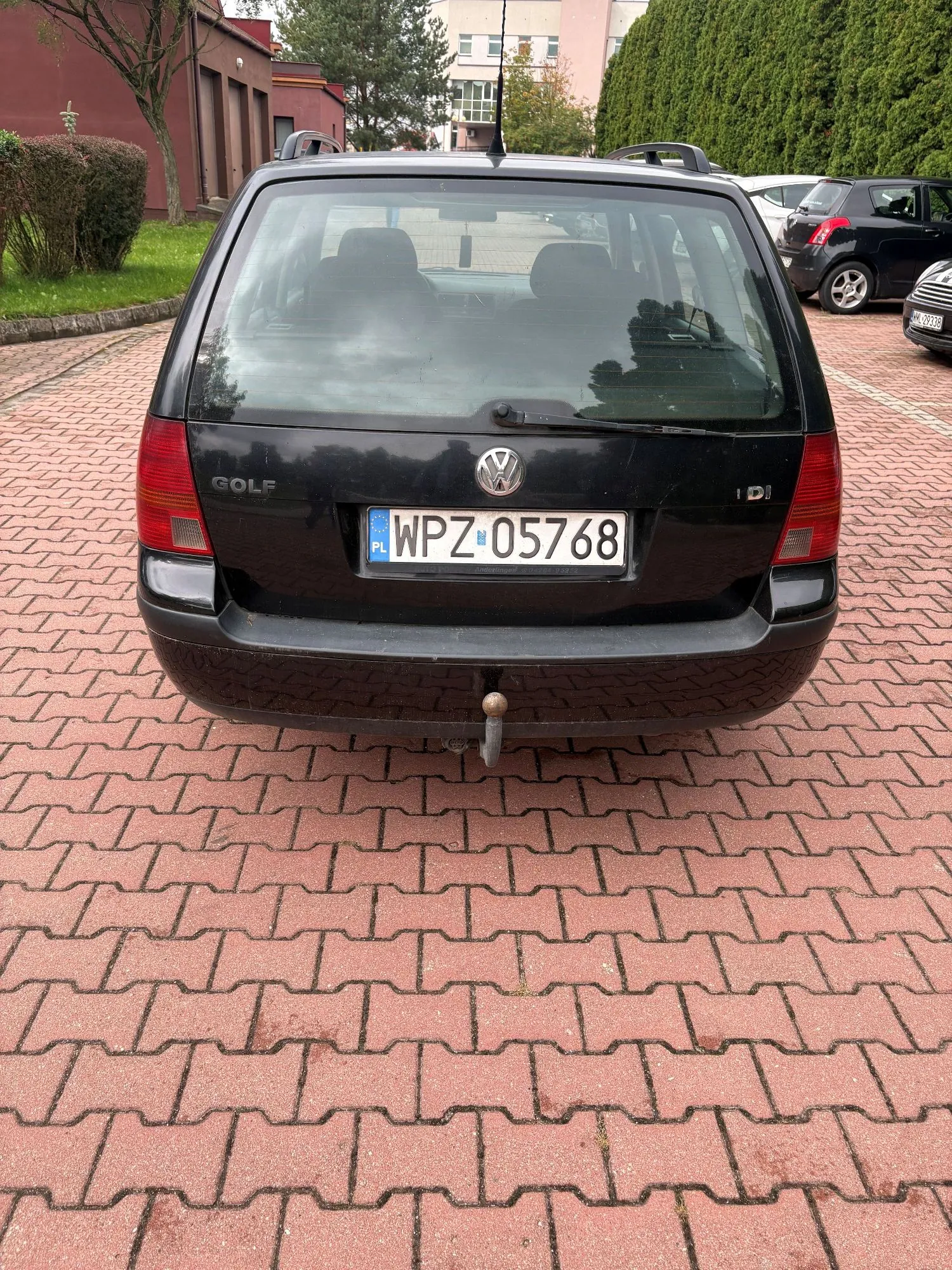 Tylny widok czarnego samochodu marki Volkswagen Golf z tablicą rejestracyjną WPZ 05768. Auto zaparkowane jest na czerwonym bruku z kostki betonowej. W tle widoczne są zielone krzewy, drzewa oraz fragmenty budynków.