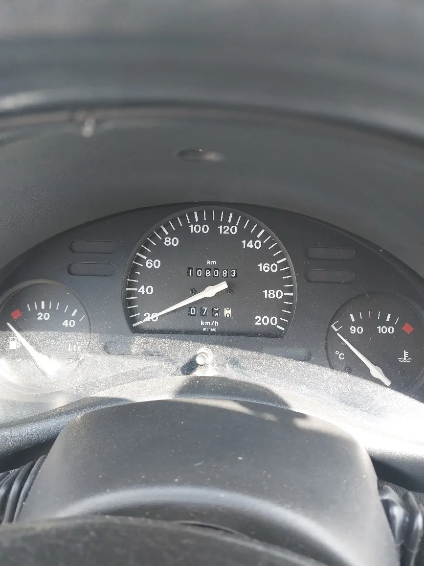 Zbliżenie na deski rozdzielcze samochodu. W centrum znajduje się prędkościomierz z zakresem do 200 km/h, wskazujący 0 km/h. Licznik przebiegu pokazuje 108083 km. Po lewej stronie zbiornik paliwa (wskaźnik blisko zera, oznaczenie Ltr), po prawej termometr chłodniczy (wskazanie ok. 90°C). Widoczna część kierownicy na dole. Tło deski rozdzielczej czarne, z białymi znakami i cyframi. Tekst na prędkościomierzu: km, km/h, W1150.