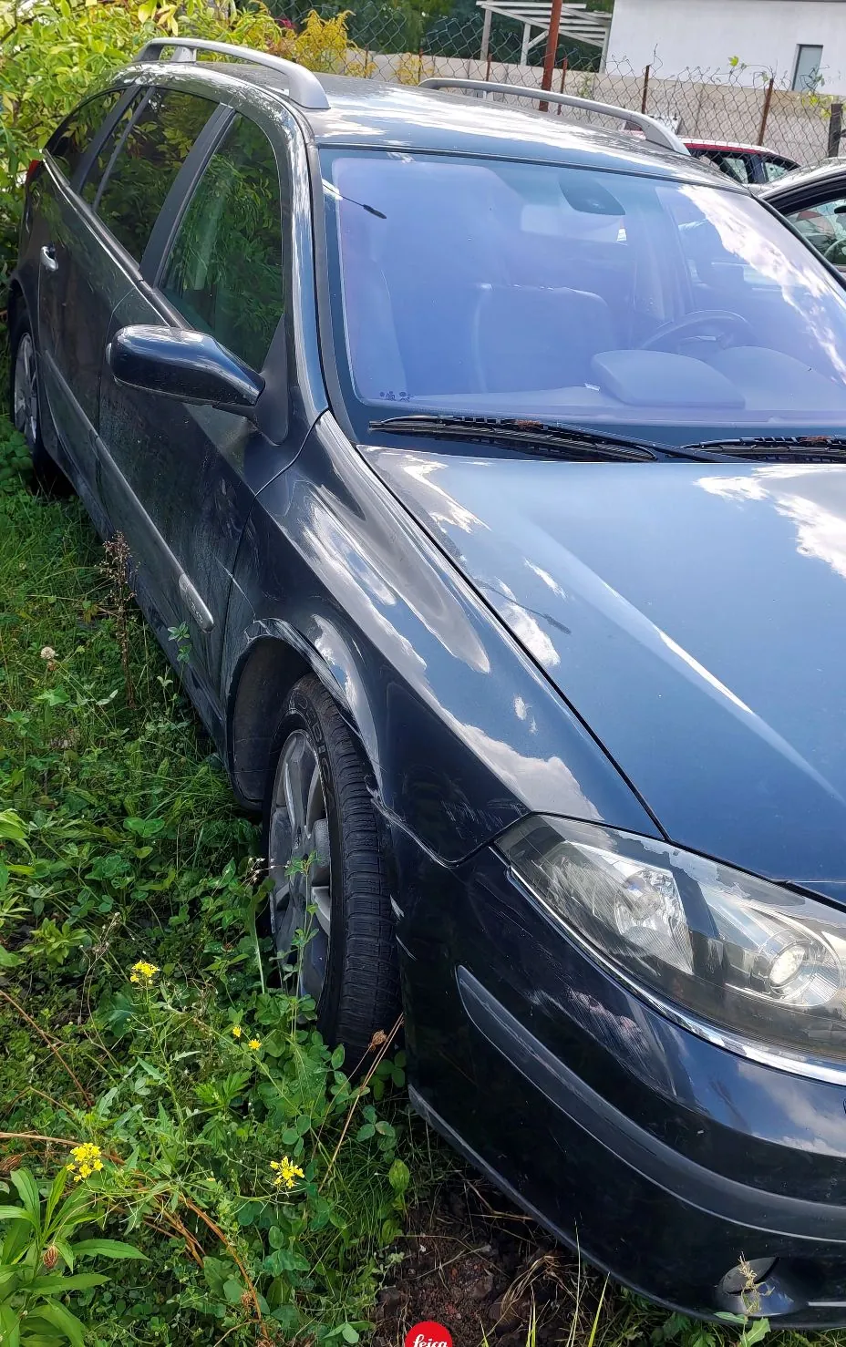 Ciemnoniebieski samochód typu kombi zaparkowany na trawiastym terenie. Widoczny jest przód i lewy bok pojazdu, w tym przednia lampa, przednia szyba z wycieraczkami oraz koło z felgą aluminiową. Na dachu znajdują się srebrne relingi. W tle widoczna jest roślinność oraz fragment białego budynku.
