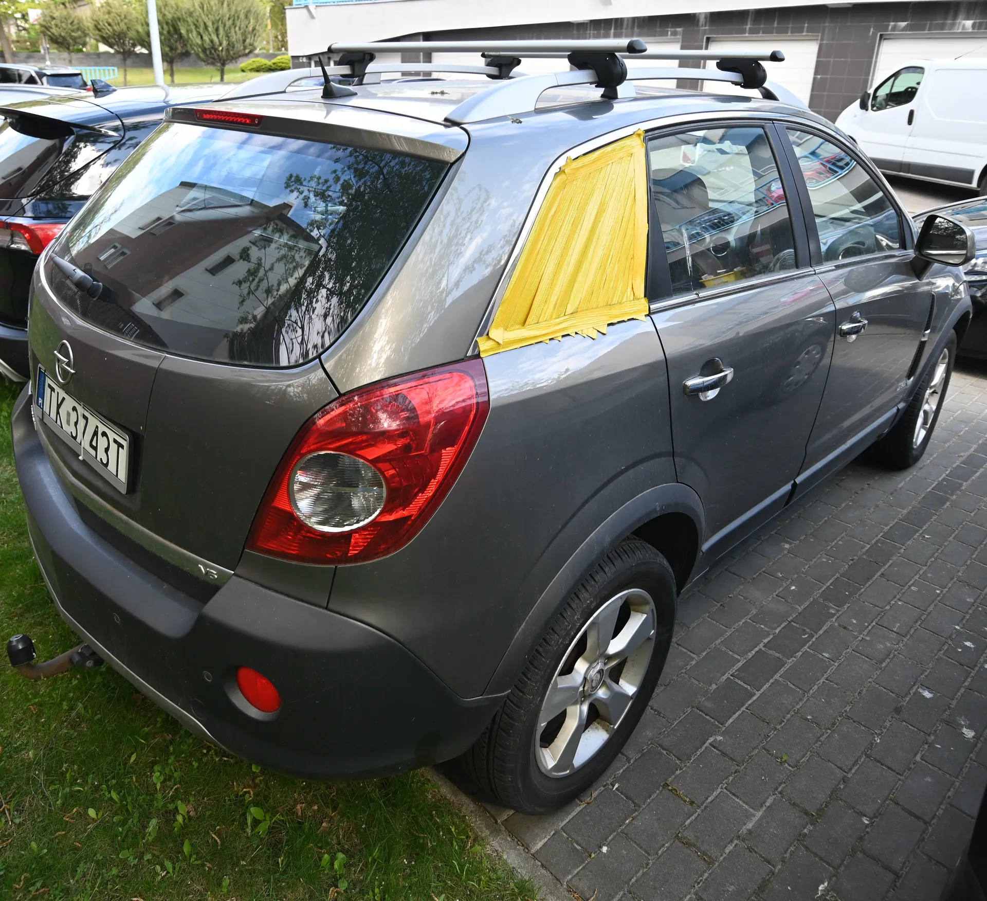 Srebrny samochód typu SUV marki Opel zaparkowany na brukowanej powierzchni. Widoczny tył pojazdu z tablicą rejestracyjną TK 3743T, czerwonymi światłami tylnymi i żółtą taśmą zakrywającą część tylnego okna po prawej stronie. Na dachu samochodu znajduje się biały dachówka. W tle widoczne są inne pojazdy, budynek z szarymi bramami garażowymi oraz zielone drzewa. Podwozie samochodu częściowo znajduje się na trawie.