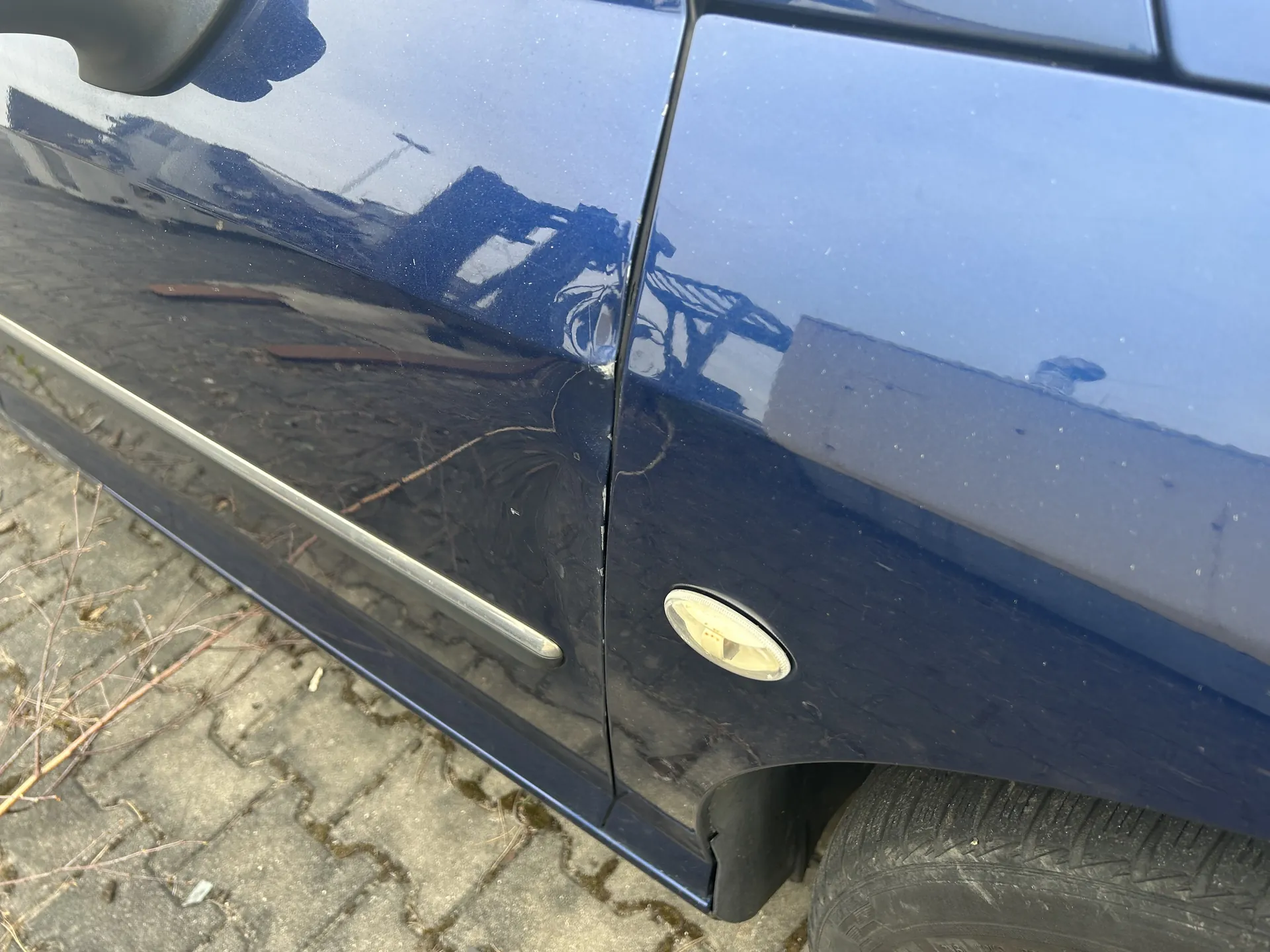 Zbliżenie na bok ciemnoniebieskiego samochodu z widoczną zadrapiną na drzwiach. Na drzwiach znajduje się srebrna rączka oraz owalny, żółtawy kierunkowskaz. W tle widoczna brukowana powierzchnia z suchymi gałązkami i mchem między kamieniami. Na błyszczącej karoserii odbijają się budynki oraz sylwetka osoby robionej zdjęcia.