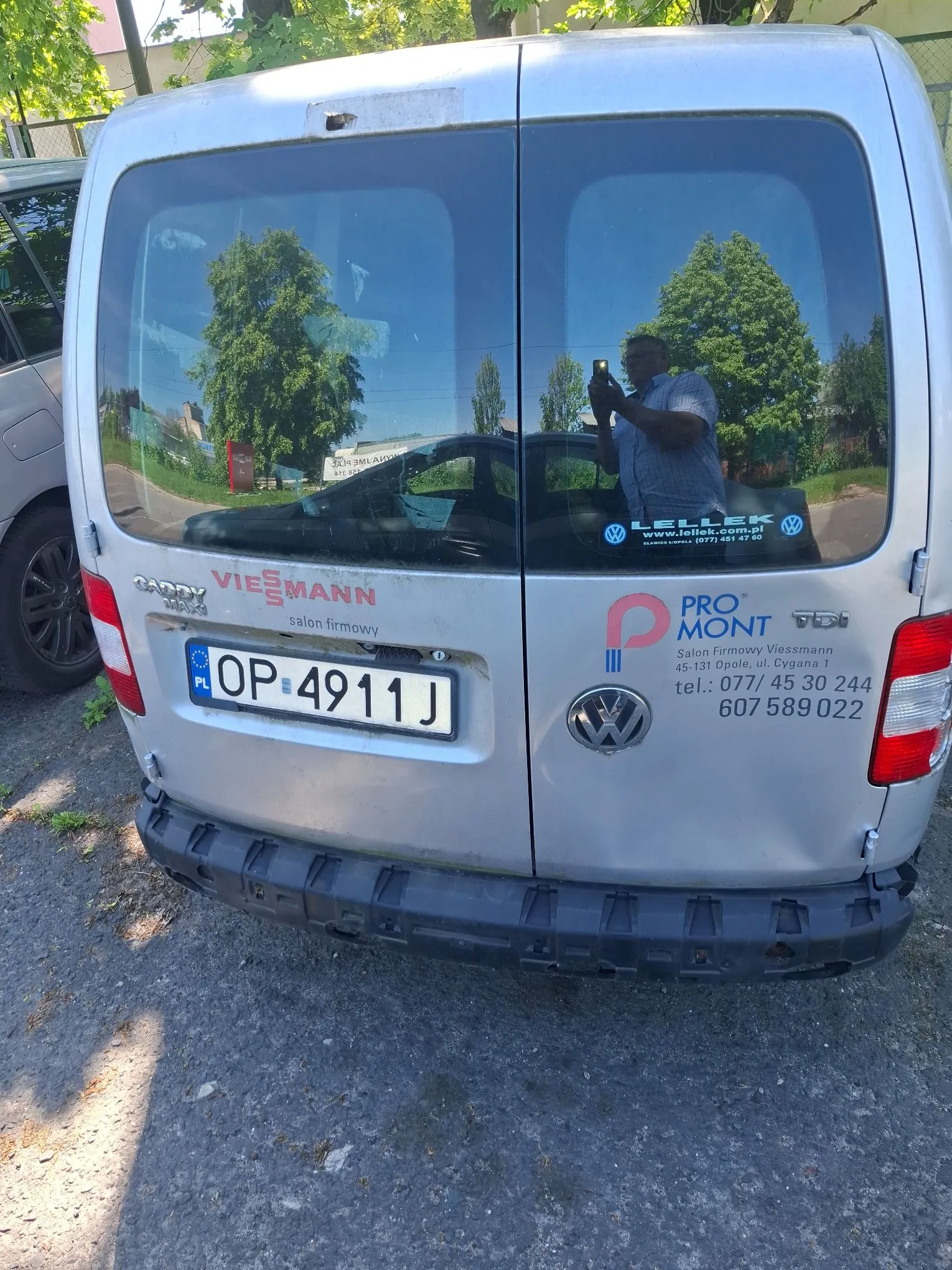Tylna część srebrnego vana marki Volkswagen. Na tylnej szybie znajdują się naklejki z napisami: 'LELLER www.lellek.com.pl', 'PROMONT' oraz dane kontaktowe: 'Salon Firmowy Viessmann 45-131 Opole, ul. Cyggana 1 tel.: 077/ 45 30 244 607 589 022'. Na klapie bagażnika widnieje logo 'VIESSMANN salon firmowy' oraz tablica rejestracyjna o numerze 'OP 4911J'. W szybie odbija się osoba robiąca zdjęcie.