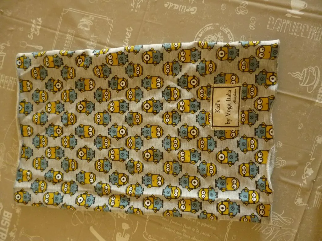 Kawałek białego materiału z nadrukiem powtarzających się, żółtych i szarych postaci przypominających Minionki. Na materiale znajduje się prostokątna, beżowa etykieta z napisem: "Kid's by Voga Italia". Materiał leży na beżowym tle z delikatnymi rysunkami kawy i napisami, takimi jak "CAPPUCCINO" i "Grillade".