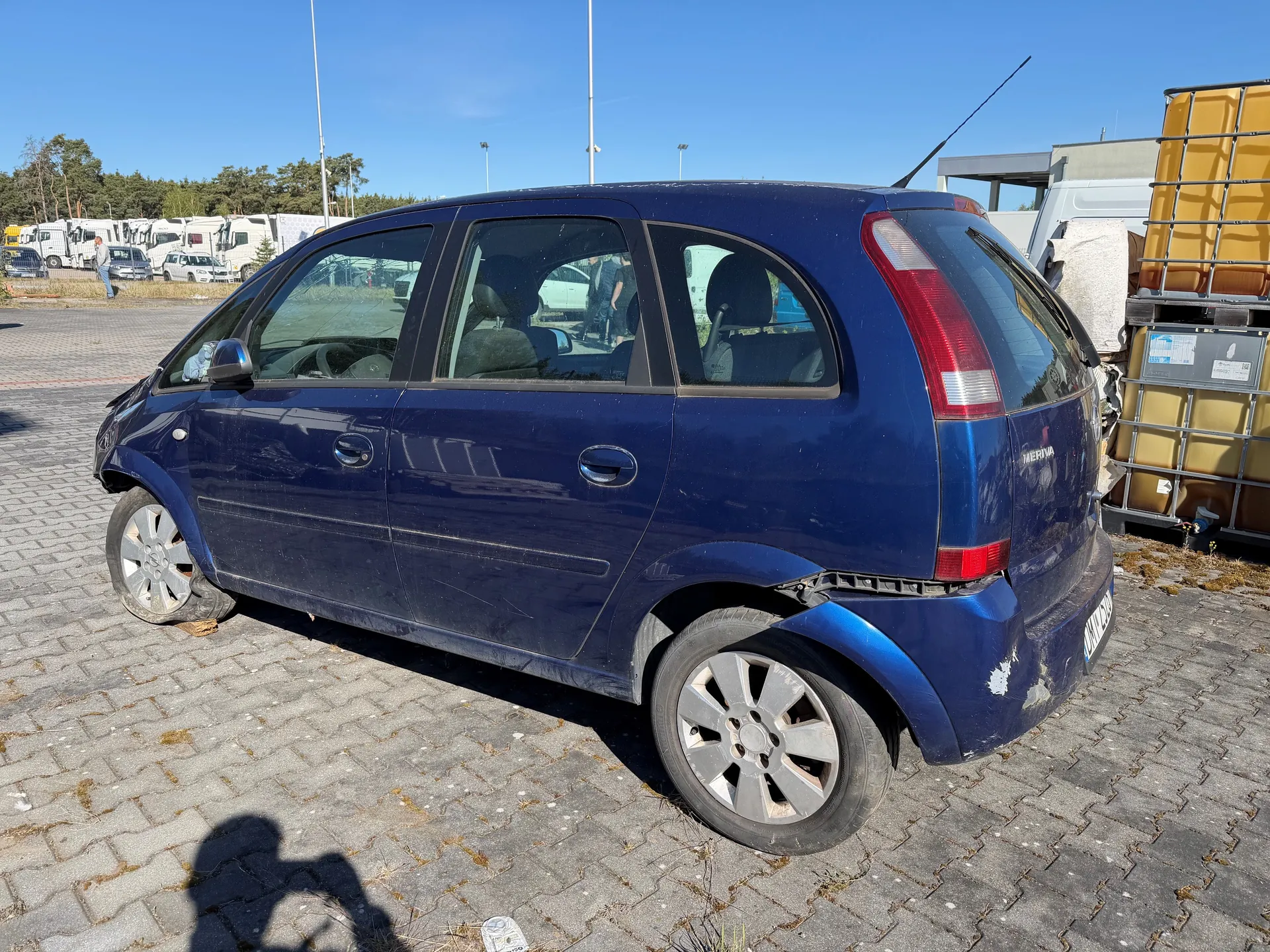 Niebieski samochód osobowy typu hatchback z widocznymi uszkodzeniami na przodzie i tylu, zaparkowany na brukowanej powierzchni. W tle widoczne są inne pojazdy, drzewa oraz żółty kontener z metalową konstrukcją. Jasne niebo bez chmur. Cień osoby fotografującej na ziemi.