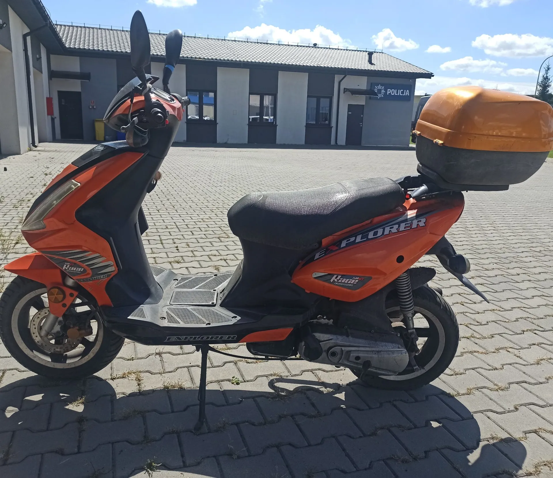 Pomarańczowy skuter marki Keeway model Explorer zaparkowany na kostce brukowej przed budynkiem policji. Skuter posiada czarne siedzenie, pomarańczowy kufer z tyłu oraz napis EXPLORER na boku. W tle widoczny jest jasny budynek z napisem POLICJA i logo policji.
