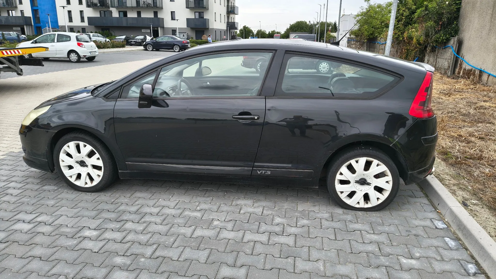 Czarny samochód osobowy marki Citroën C4 Coupe zaparkowany bokiem na kostce brukowej. Na dolnej części drzwi widoczny jest napis 'VTS'. W tle znajdują się białe samochody, nowoczesny budynek mieszkalny oraz zieleń.
