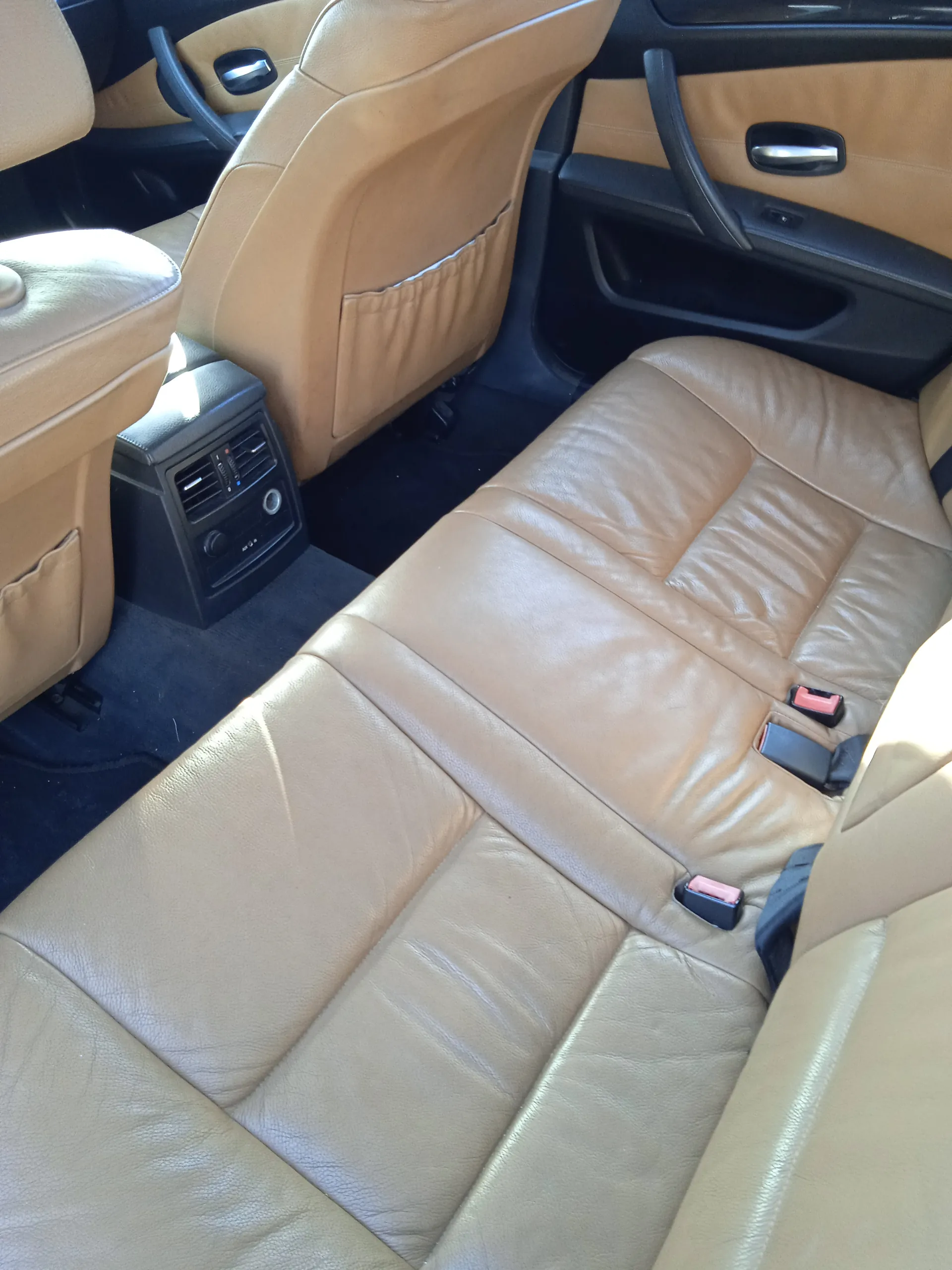 Wnętrze samochodu z widokiem na tylną część kabiny. Beżowe skórzane siedzenia z widocznymi pasami bezpieczeństwa o czerwonych i czarnych zapięciach. Widoczne są również przednie siedzenia, czarna konsoleta środkowa z wentylacją i przyciskami oraz drzwi z beżowymi panelami i czarnymi uchwytami. Podłoga wyłożona czarnym dywanem.
