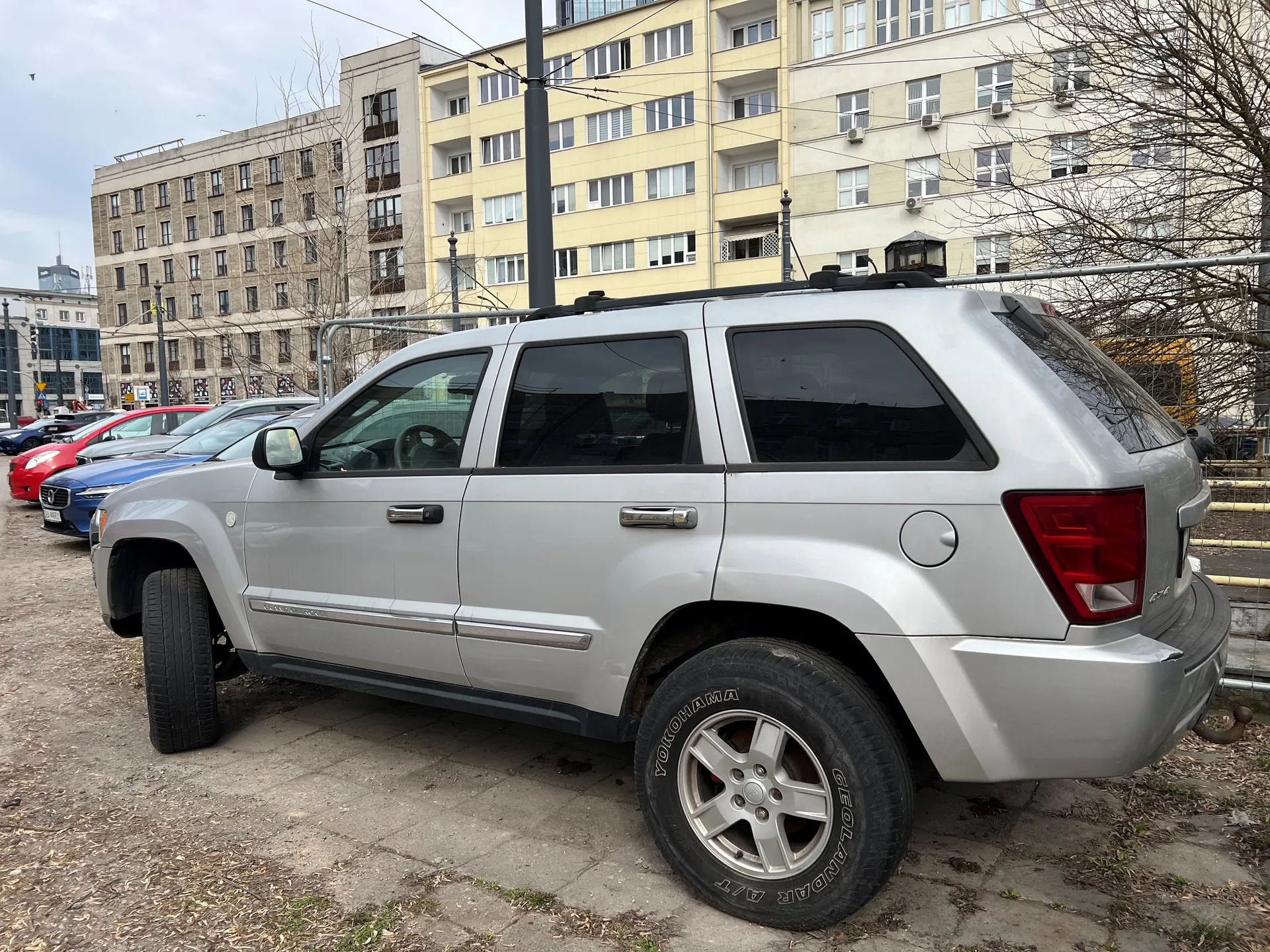Srebrny SUV zaparkowany na otwartym parkingu. W tle widoczne są inne samochody (czerwony, niebieski), wielopiętrowe budynki mieszkalne z oknami i drzewa bez liści. Na oponie tylnego koła widoczny napis YOKOHAMA GEOLANDAR A/T. Tło stanowi chmurne niebo i fragmenty innych budynków.
