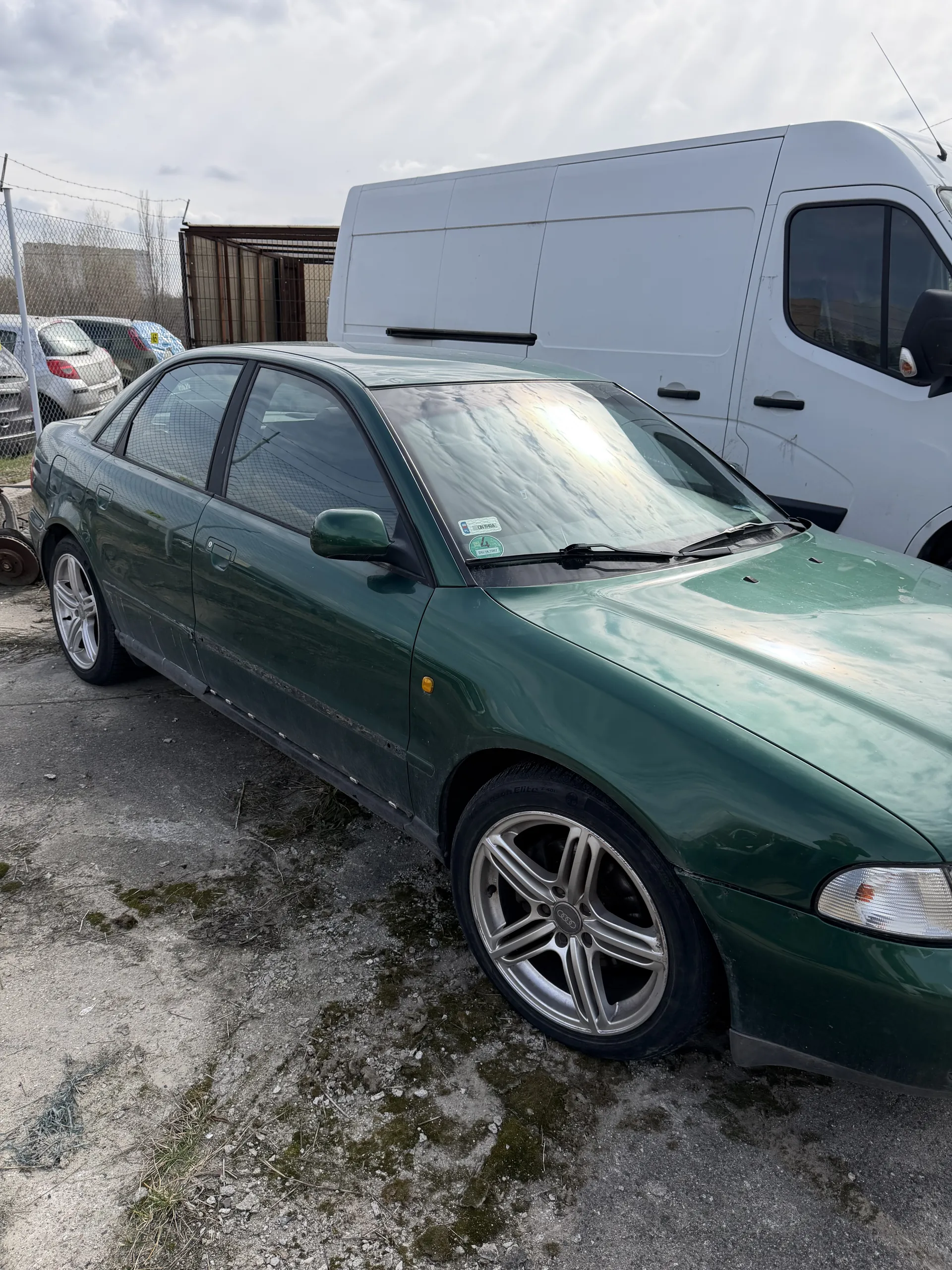 Zielony samochód osobowy marki Audi z srebrnymi felgami, zaparkowany obok białego furgonetki. W tle widoczne są inne pojazdy, ogrodzenie siatkowe, budynek z metalową konstrukcją oraz chmurne niebo. Podłoga z żwiru i mchu. Na przedniej szybie zielonego samochodu znajdują się naklejki.