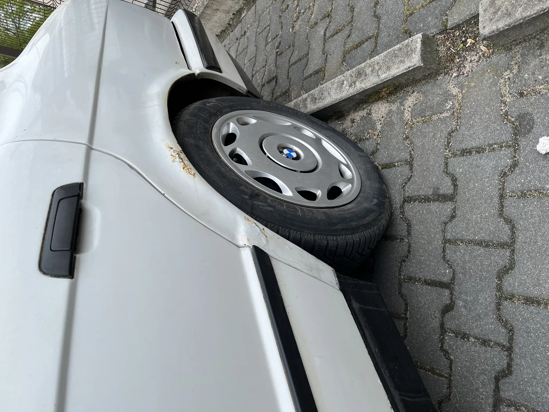 Biały samochód z przodem na krawężniku, koło przednie na chodniku. Widoczny srebrny felg z logo BMW w centrum koła. Na nadkolu i nadwoziu ślady rdzy. Tło: brukowana droga z kamiennymi płytami, betonowy krawężnik oraz metalowe ogrodzenie z zielonymi elementami w tle.