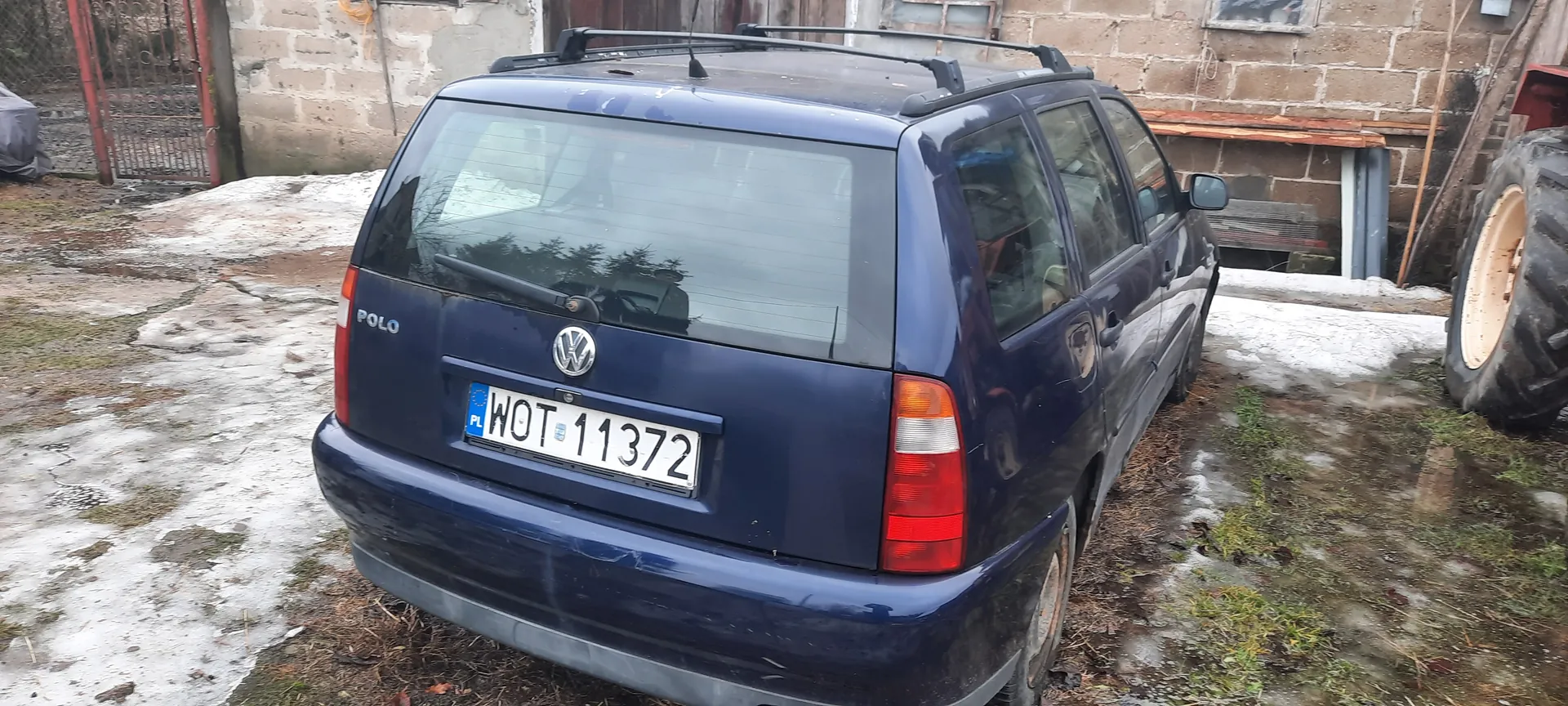Ciemnoniebieski Volkswagen Polo z napisem "POLO" i logo VW na tylnej szybie. Tablica rejestracyjna PL WOT 11372. Samochód z bagażnikiem na dachu zaparkowany na błotnistym podłożu z resztkami śniegu. W tle ceglany budynek, brama i fragment opony traktora.