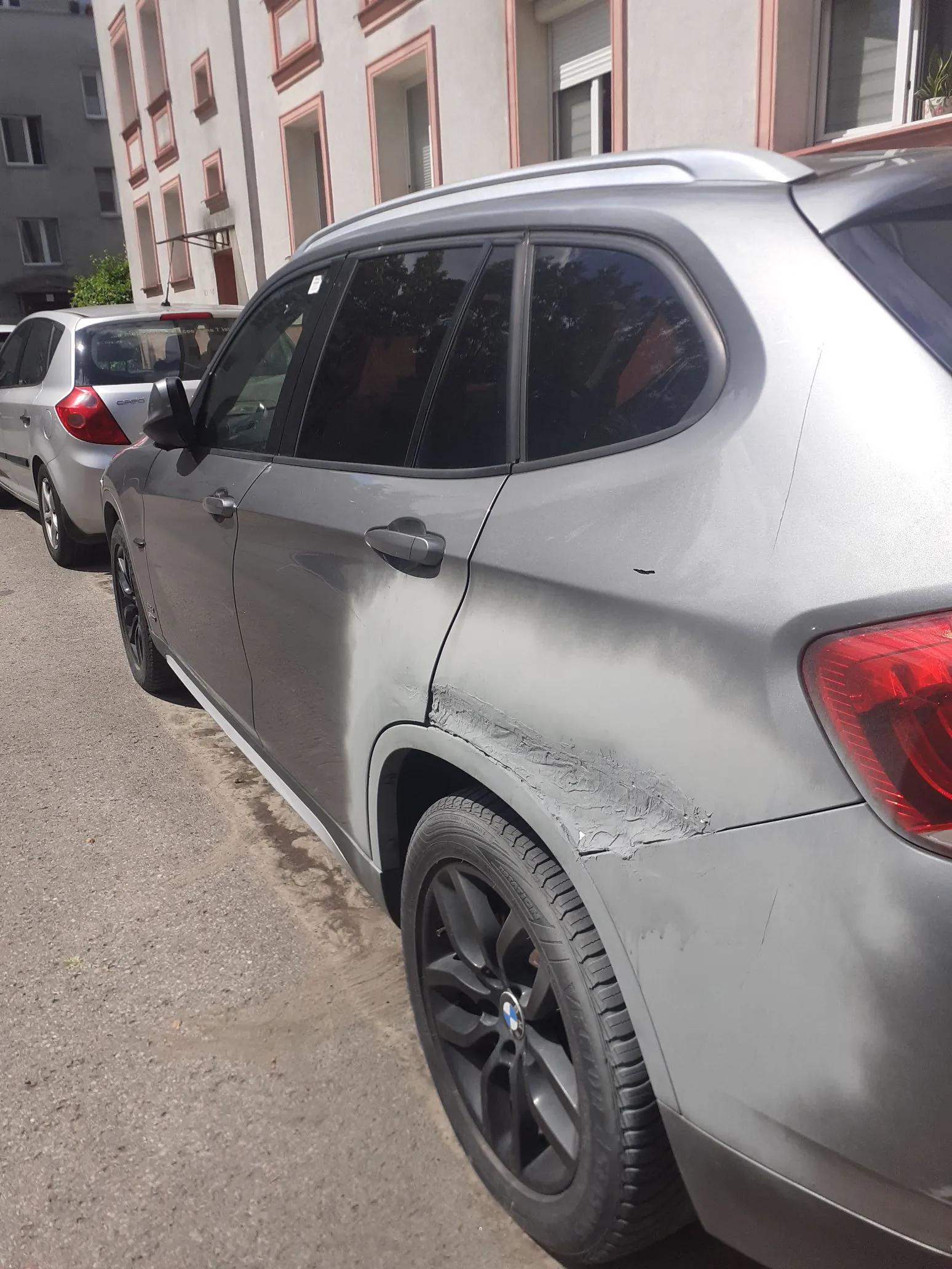 Zbliżenie na prawy bok srebrnego samochodu typu SUV. Na tylnym błotniku, nad kołem, widoczne są uszkodzenia karoserii w postaci wgnieceń oraz śladów naprawy szpachlowej, która odpryskuje. Samochód zaparkowany jest przy krawężniku obok jasnego budynku mieszkalnego. W tle widoczny jest fragment innego, białego samochodu.
