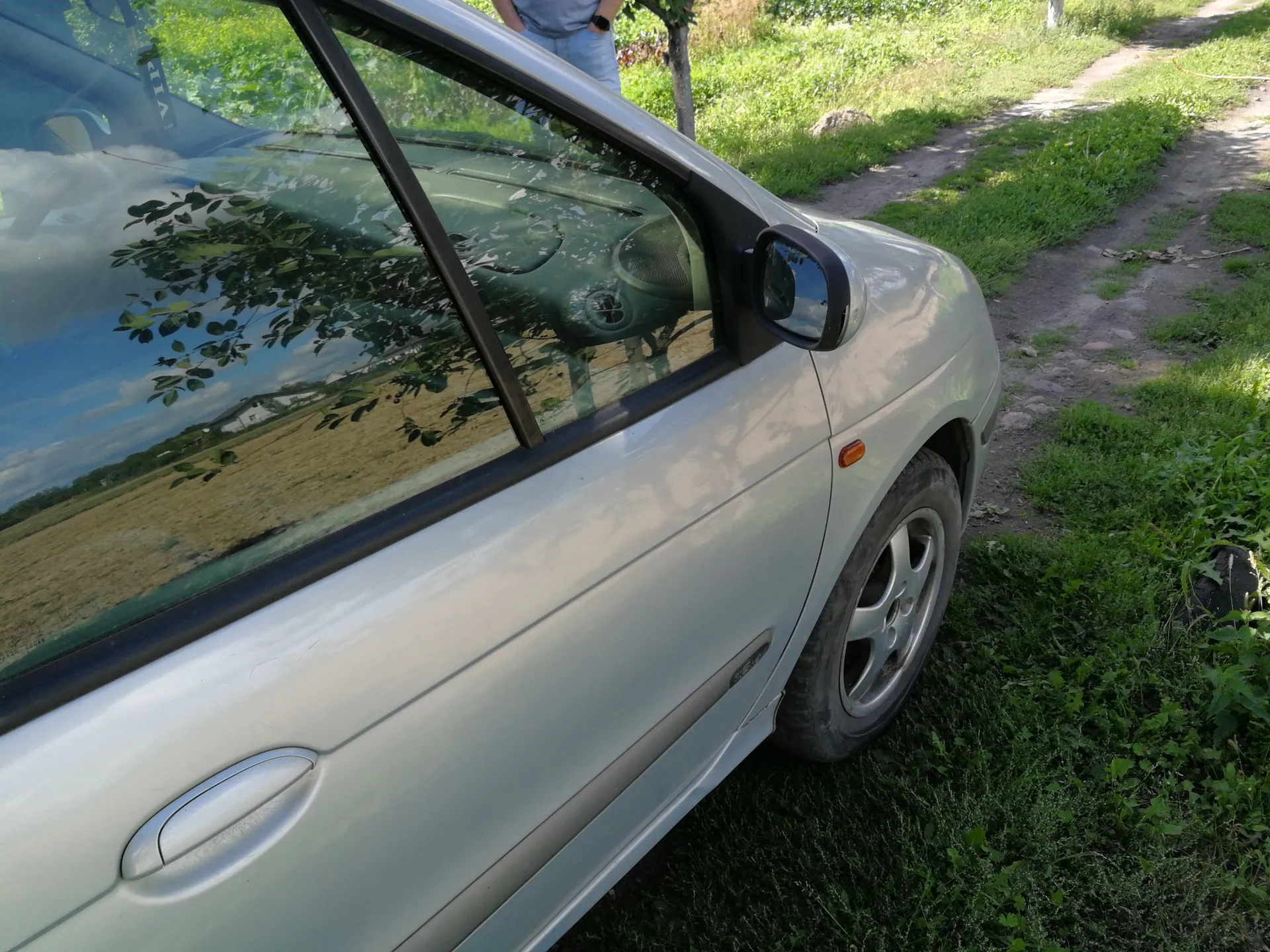 Zbliżenie na srebrny samochód osobowy zaparkowany na trawiastej poboczu obok polnej drogi. Widoczny jest fragment przednich drzwi, lusterko boczne oraz przednie koło z felgą aluminiową. W szybach samochodu odbija się niebo, drzewa i okoliczny krajobraz.