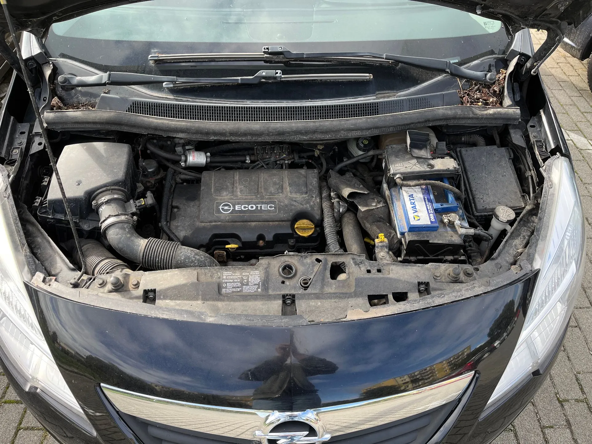 Widok z góry na otwartą maskę samochodu, ukazującą komorę silnika. W centrum znajduje się czarna osłona silnika z białym napisem 'ECOTEC' i logo marki Opel. Po prawej stronie widoczny jest akumulator marki Varta w niebiesko-białej obudowie. Po lewej stronie znajduje się obudowa filtra powietrza i rury dolotowe. Elementy są zakurzone.