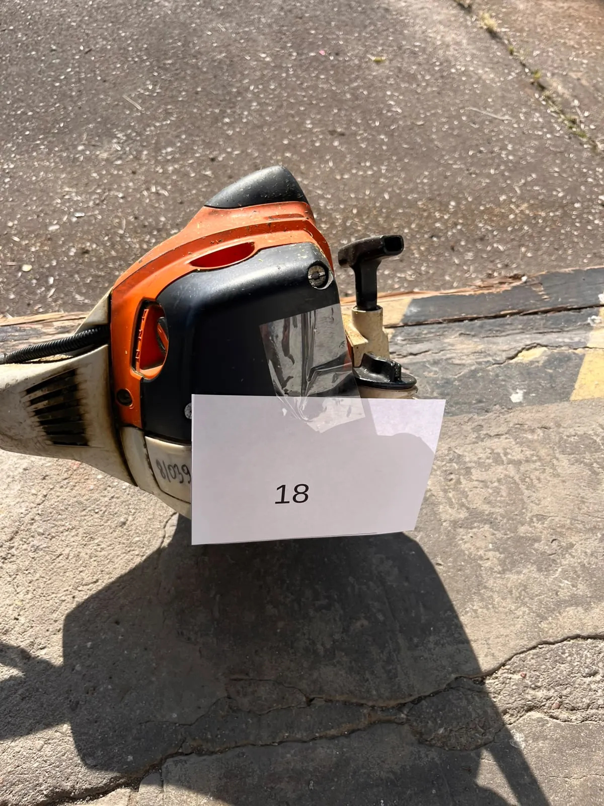 Pomarańczowo-czarna piła spalinowa leżąca na szarym, betonowym podłożu. Na urządzeniu przyklejona jest biała kartka z czarnym napisem „18”. Na obudowie piły widoczny jest również odręczny numer „81039”.
