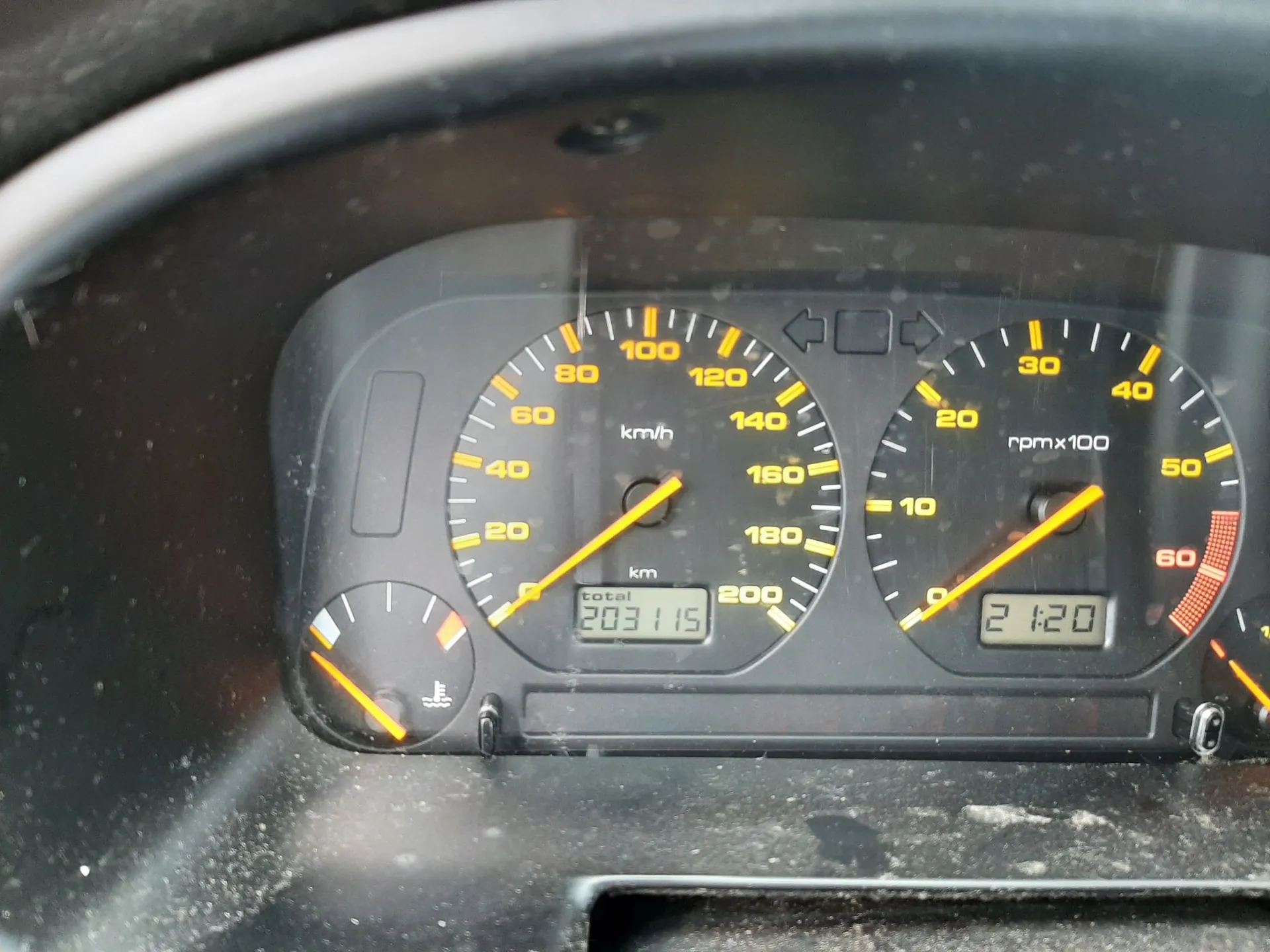 Zbliżenie na deskę rozdzielczą samochodu. Widoczny jest prędkościomierz z zakresem do 200 km/h, obrotomierz z oznaczeniem rpm x 100 oraz licznik kilometrów z wartością 203115 km. Poniżej obrotomierza znajduje się zegar cyfrowy wskazujący godzinę 21:20. Po lewej stronie widoczny jest wskaźnik poziomu paliwa.