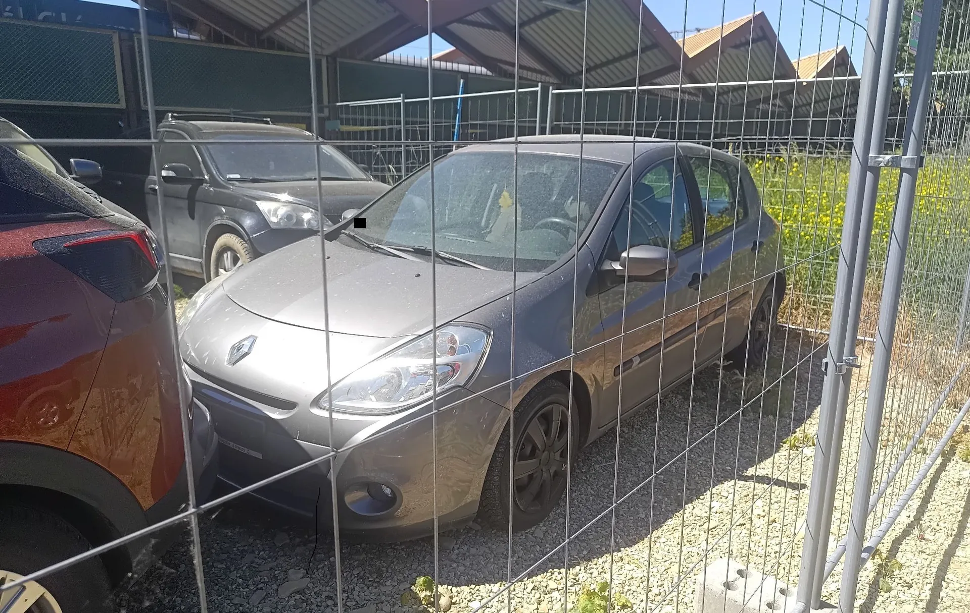 Zdjęcie wykonane przez metalowy płot z siatki, przedstawiające zaparkowane samochody. Na pierwszym planie widać fragment czerwonego auta po lewej stronie oraz szary samochód osobowy marki Renault w centrum. W tle znajduje się kolejny, ciemnoszary samochód oraz zarysy budynków z trójkątnymi dachami pod jasnym, słonecznym niebem.