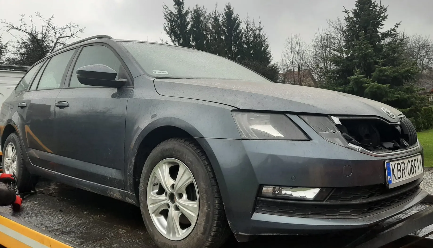 Szary samochód osobowy typu kombi marki Škoda z uszkodzonym przodem, stojący na platformie holownika. Widoczna tablica rejestracyjna KBR-08911 z oznaczeniem PL. Samochód jest zabrudzony, a na tle znajdują się drzewa i pochmurne niebo. W lewym dolnym rogu widoczna część ręki w czerwonej rękawicy.