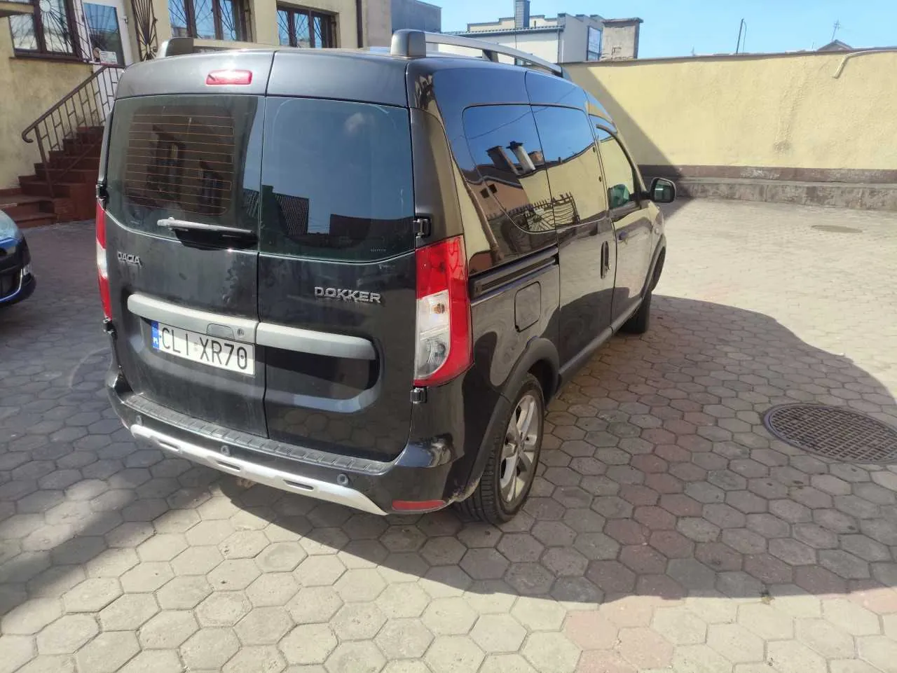 Czarny samochód dostawczy Dacia Dokker zaparkowany na brukowanej powierzchni. Widok od tyłu z tablicą rejestracyjną CL1 XR70. W tle budynki, schody z metalowym poręczem oraz jasne niebo.