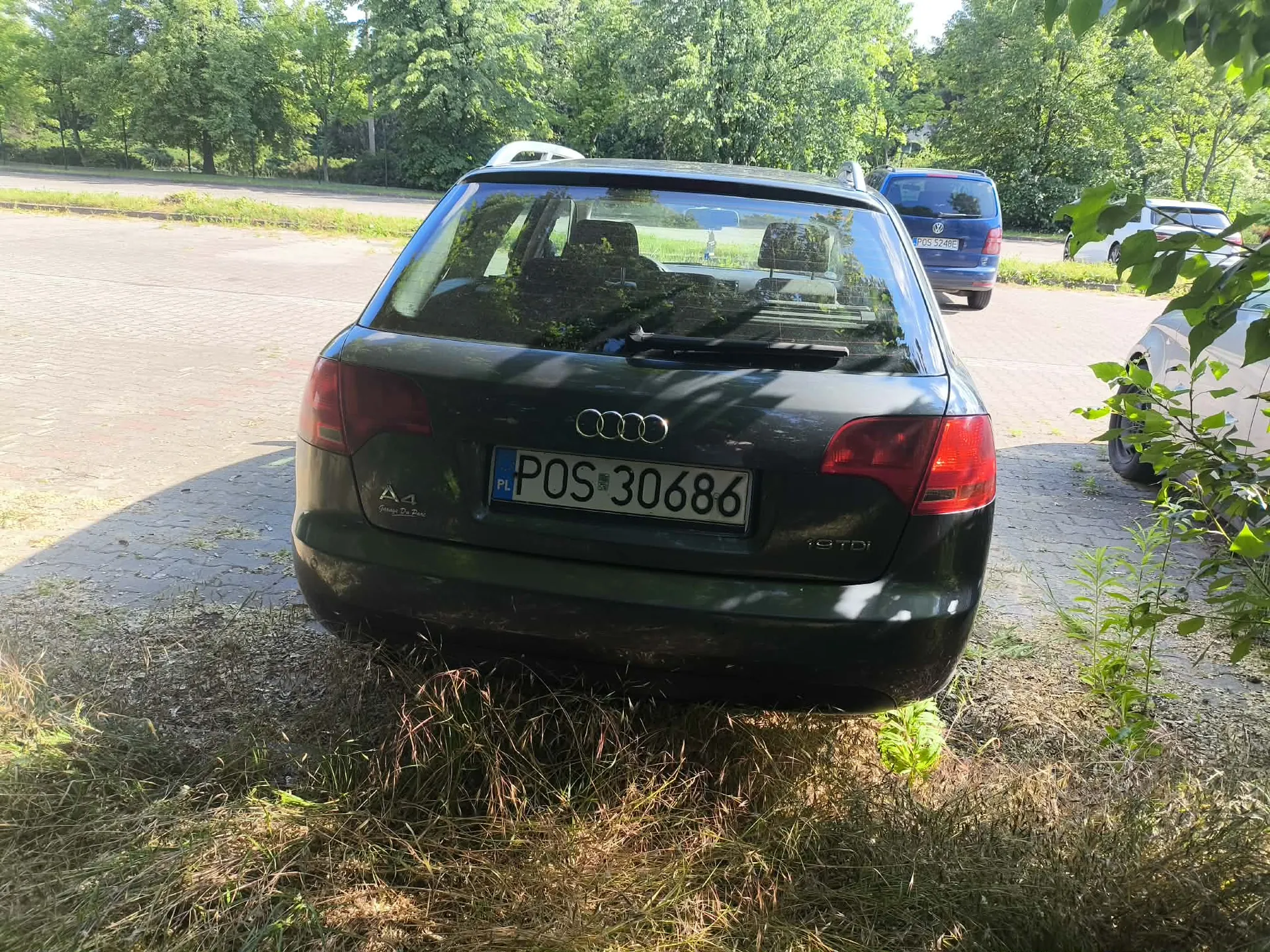 Tylny widok ciemnozielonego samochodu marki Audi A4 zaparkowanego na utwardzonym placu obok trawy. Na klapie bagażnika widoczne jest logo Audi oraz tablica rejestracyjna o numerze POS 30686. Po lewej stronie znajduje się oznaczenie modelu A4, a po prawej napis TDI. W tle widoczne są inne zaparkowane samochody oraz gęste, zielone drzewa.
