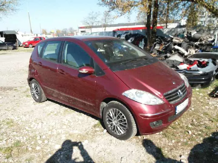 Czerwony samochód osobowy marki Mercedes-Benz klasy B zaparkowany na żwirowym placu. W tle widoczne są zniszczone pojazdy, fragment budynku z czerwono-białą pasą oraz drzewa. Na pierwszym planie cienie osób i nierówna powierzchnia ziemi z rzadką trawą.